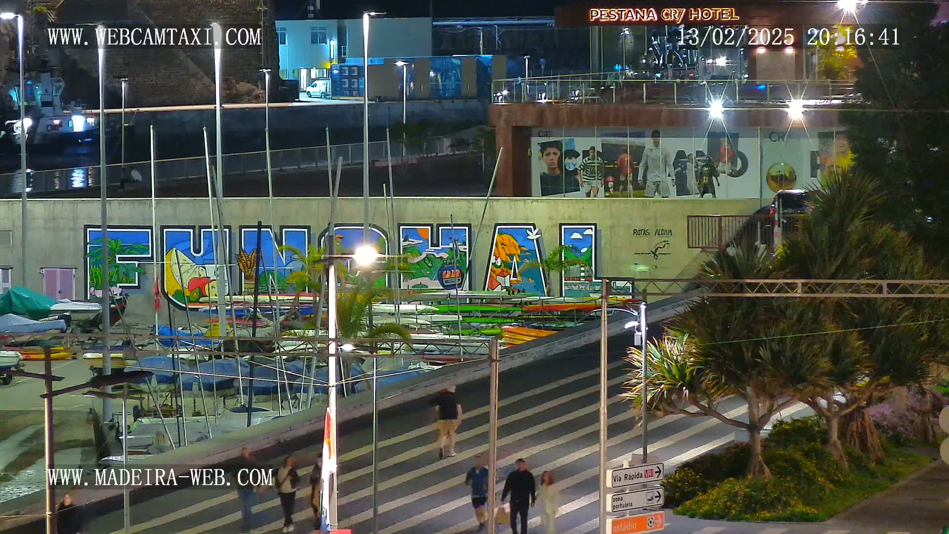 A nighttime harbor scene shows a large mural of the word "Funchal" on a building, with numerous boats and kayaks docked below, and pedestrians walking along a striped walkway.