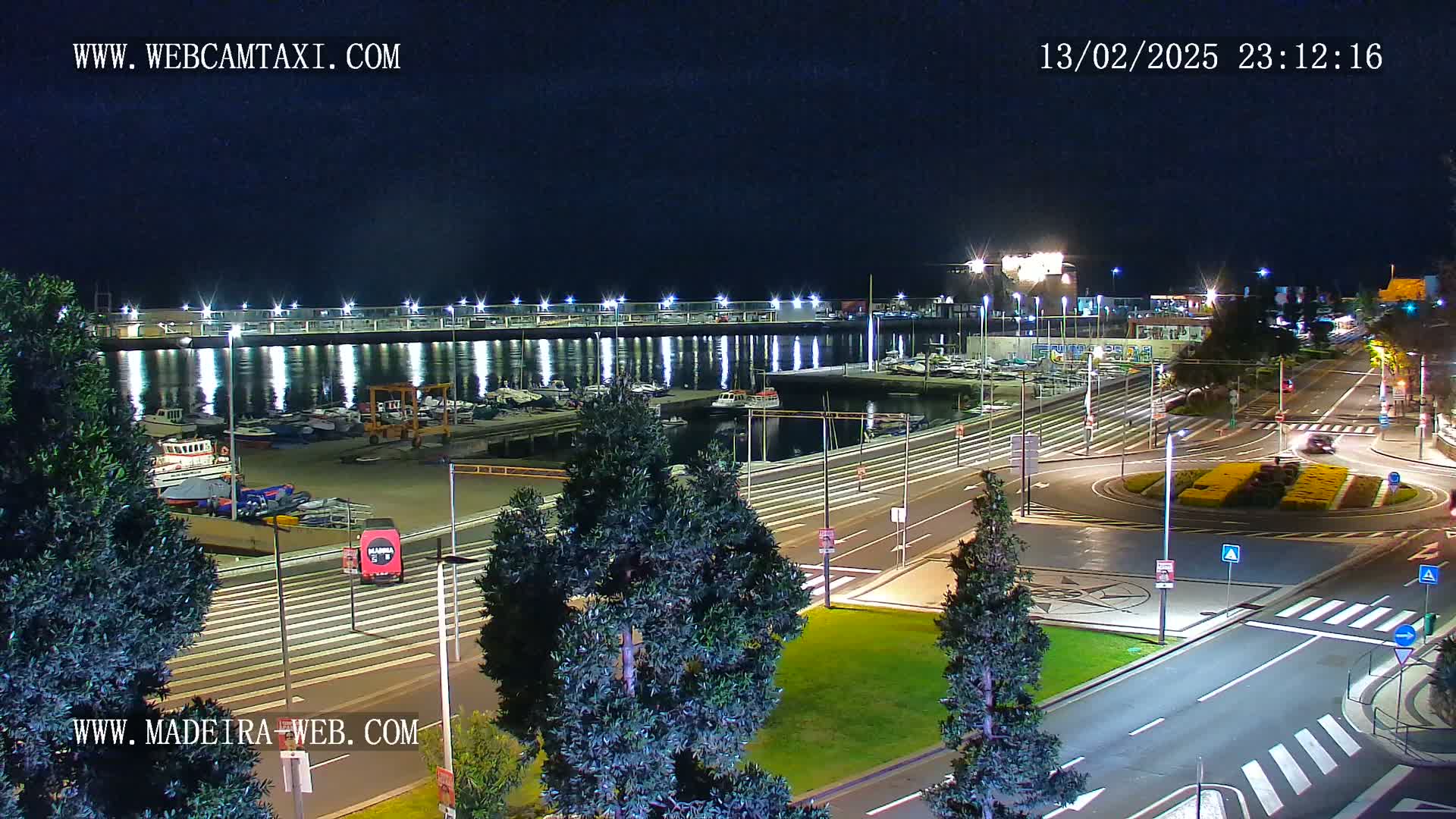 A nighttime view of a harbor with boats docked, illuminated by lights, next to a roadway with a roundabout, under a clear sky.