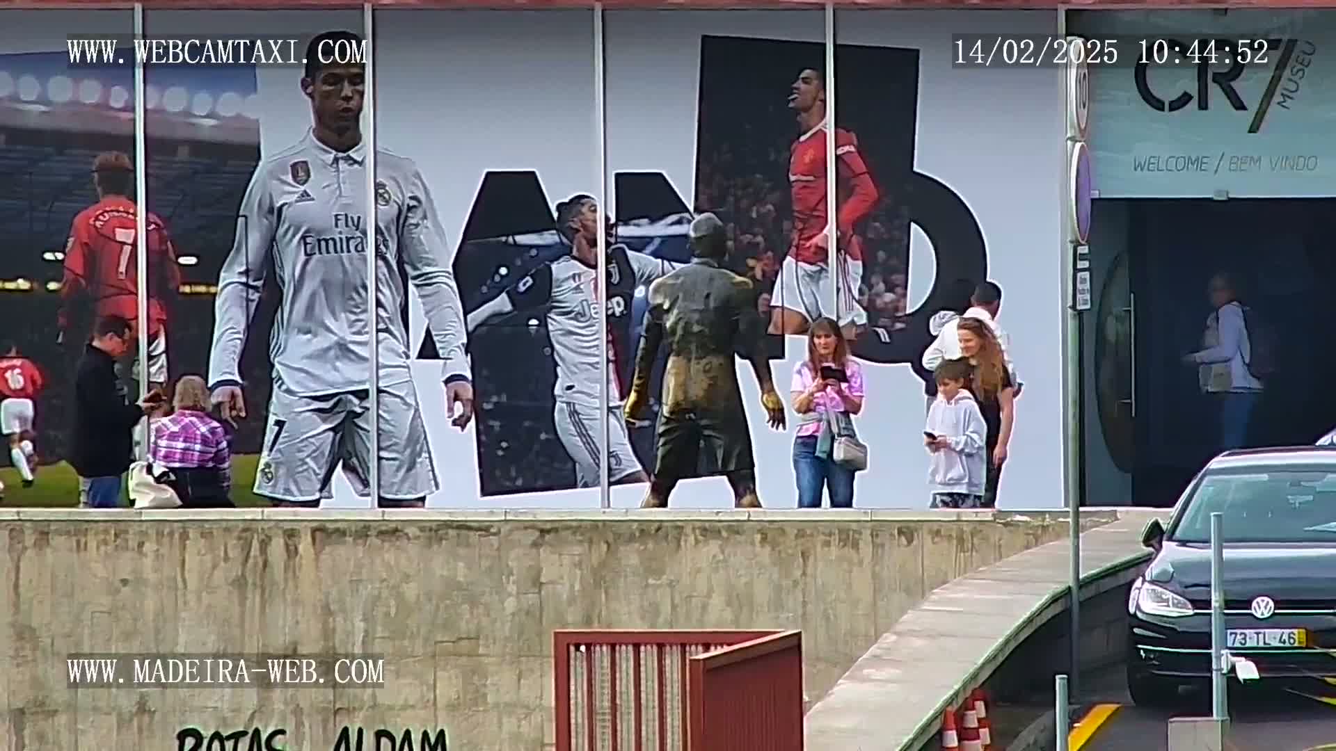A large billboard depicting Cristiano Ronaldo in various soccer jerseys is displayed outside a building, with several people standing in front of it on a sunny day.