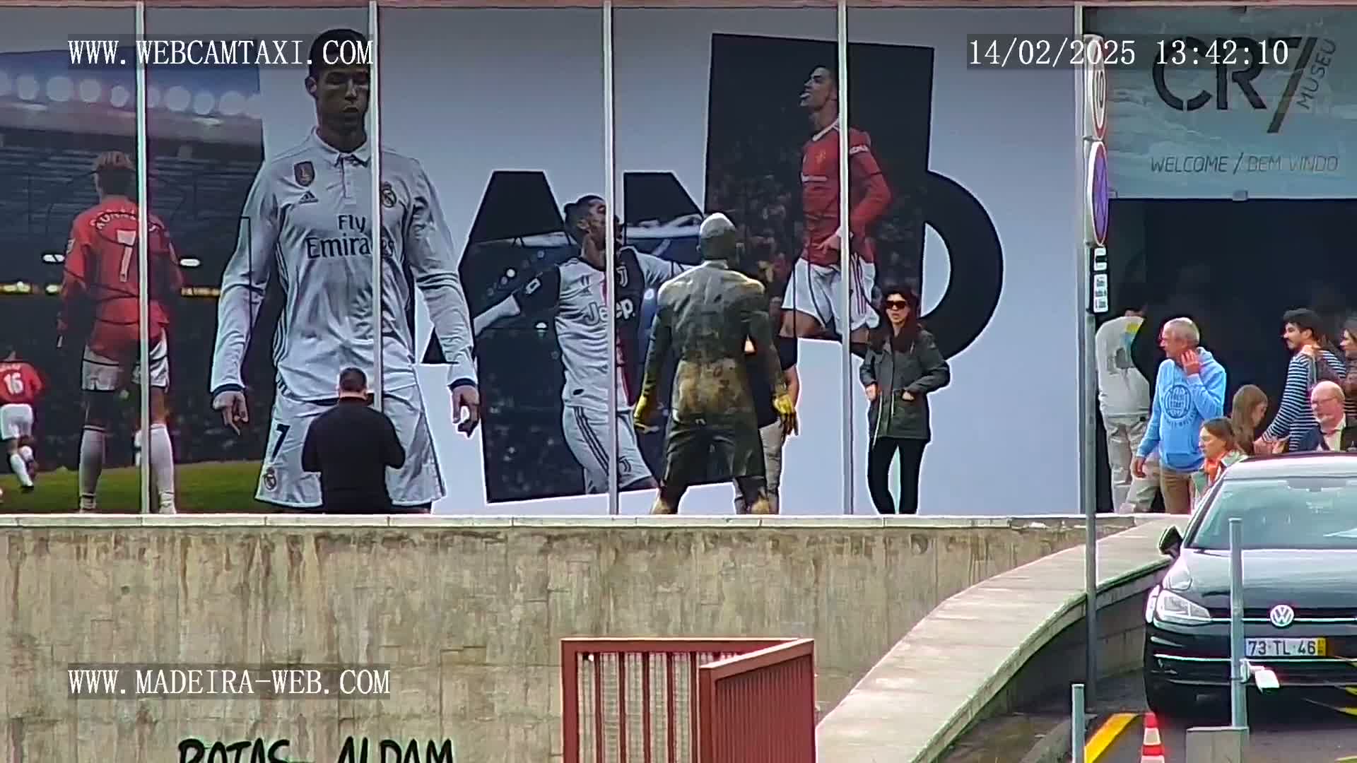 A large outdoor mural depicting Cristiano Ronaldo and other soccer players is viewed from across a short wall, with several people standing near a car in front of it on a somewhat overcast day.