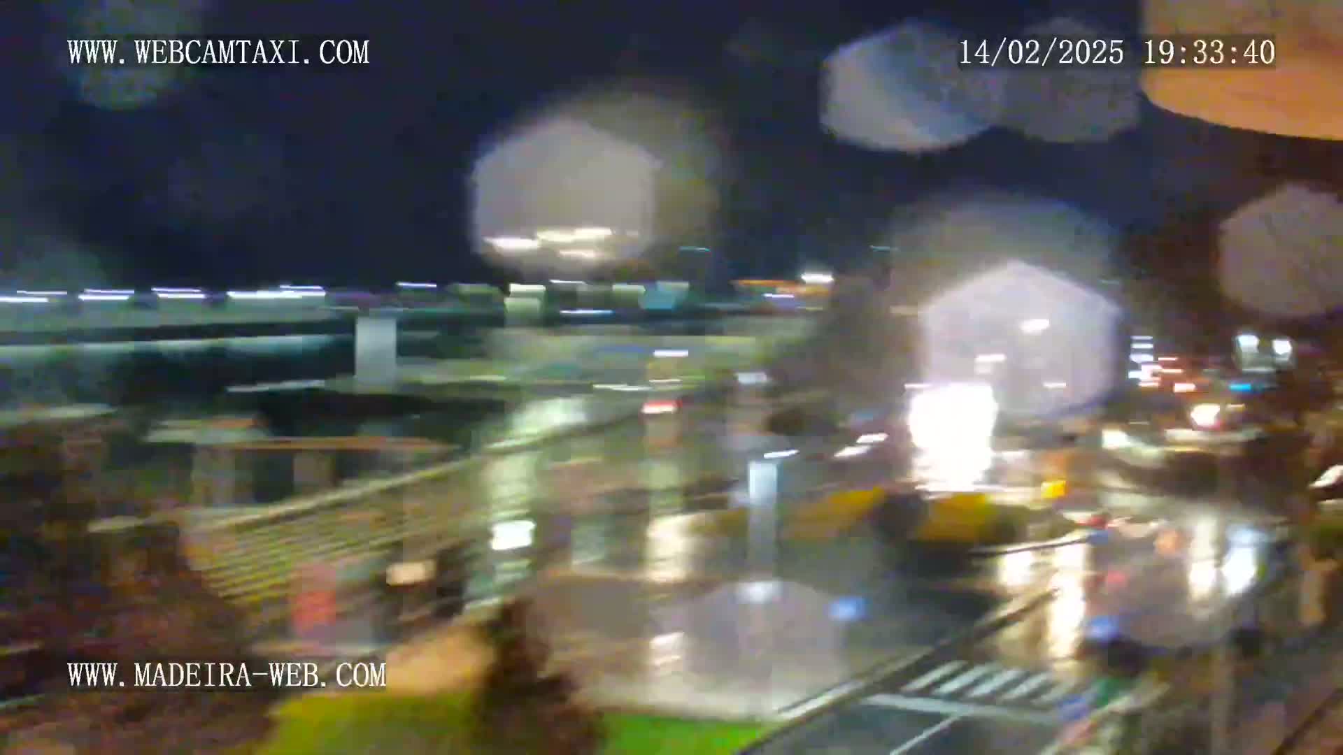 A blurry nighttime view of a rain-slicked city street scene, showing lights and blurred vehicles through a rain-covered window.