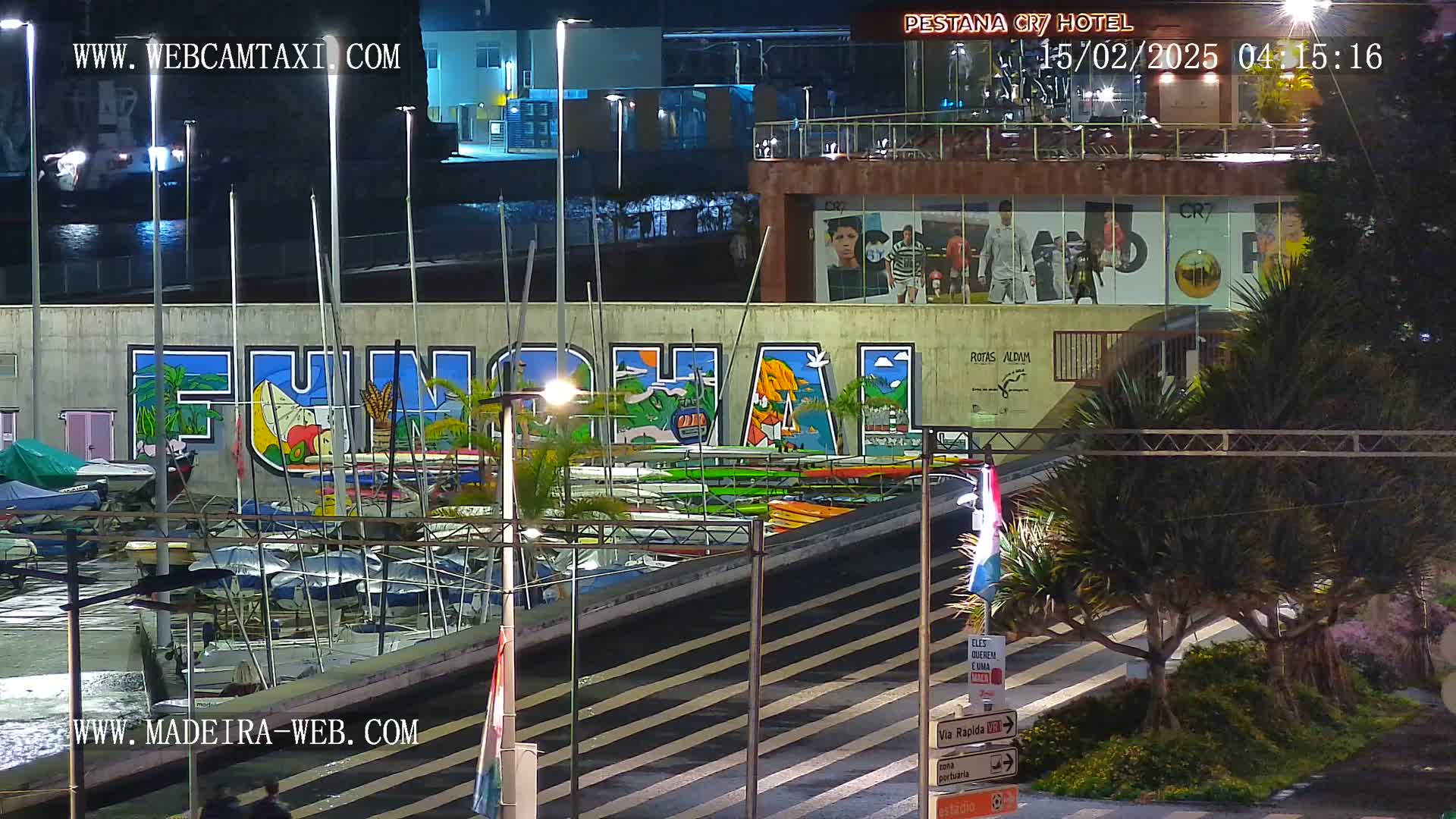 At night, a marina with numerous boats is shown in front of a wall with a large mural and a building with large advertisements.