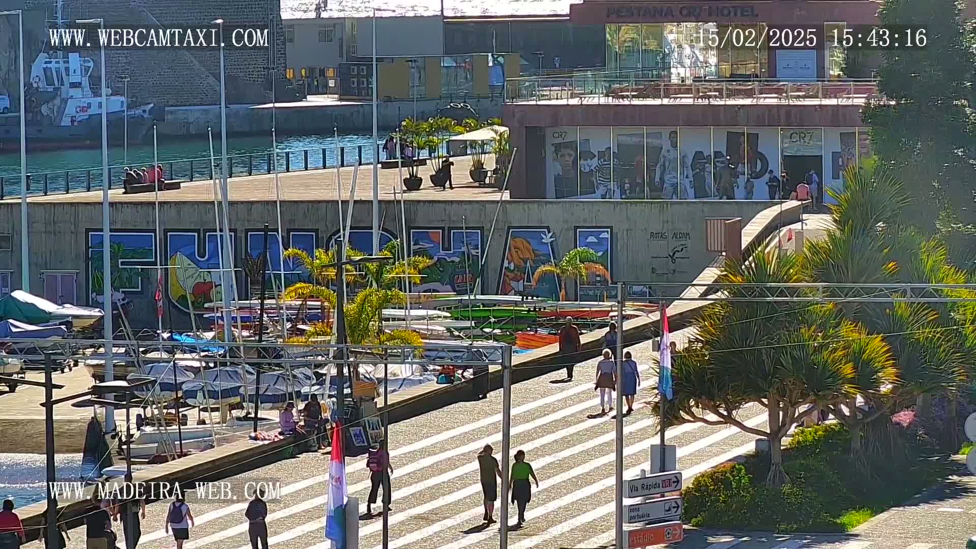 A sunny harbor scene shows numerous sailboats docked alongside a building with colorful graffiti, and pedestrians walking along a paved walkway.