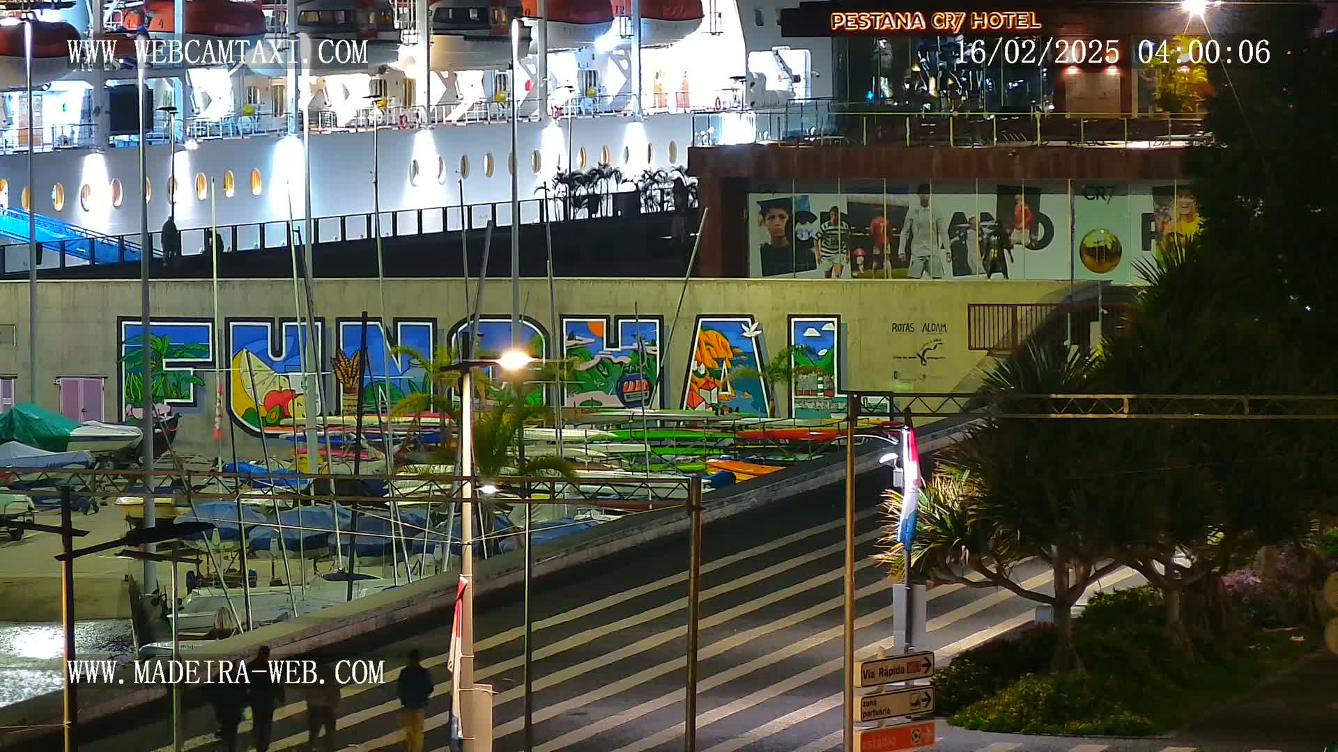 A nighttime harbor scene shows a large ship docked next to a building with colorful murals, numerous small boats at the water's edge, and palm trees under streetlights.