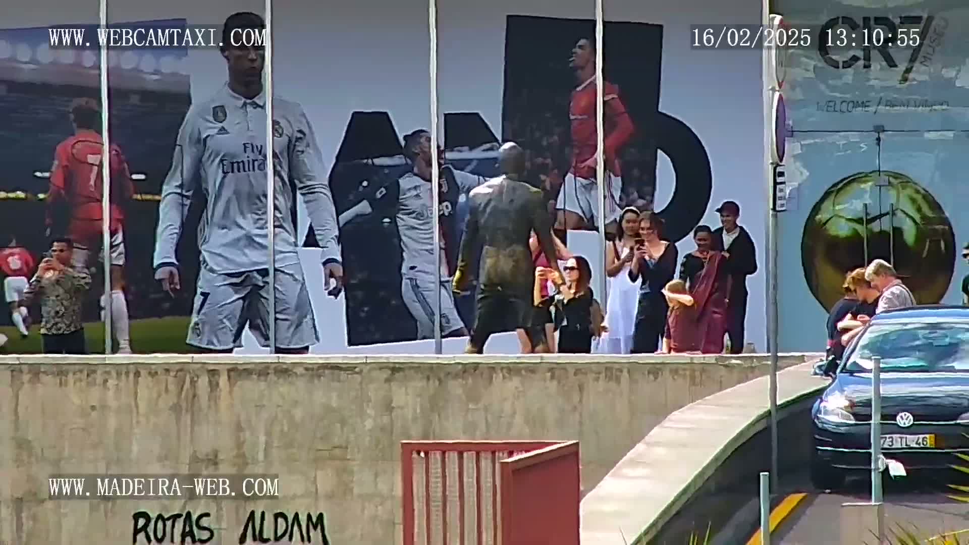A large billboard depicting soccer players is viewed from across a street, where several people are standing and a car is parked, on a sunny day.