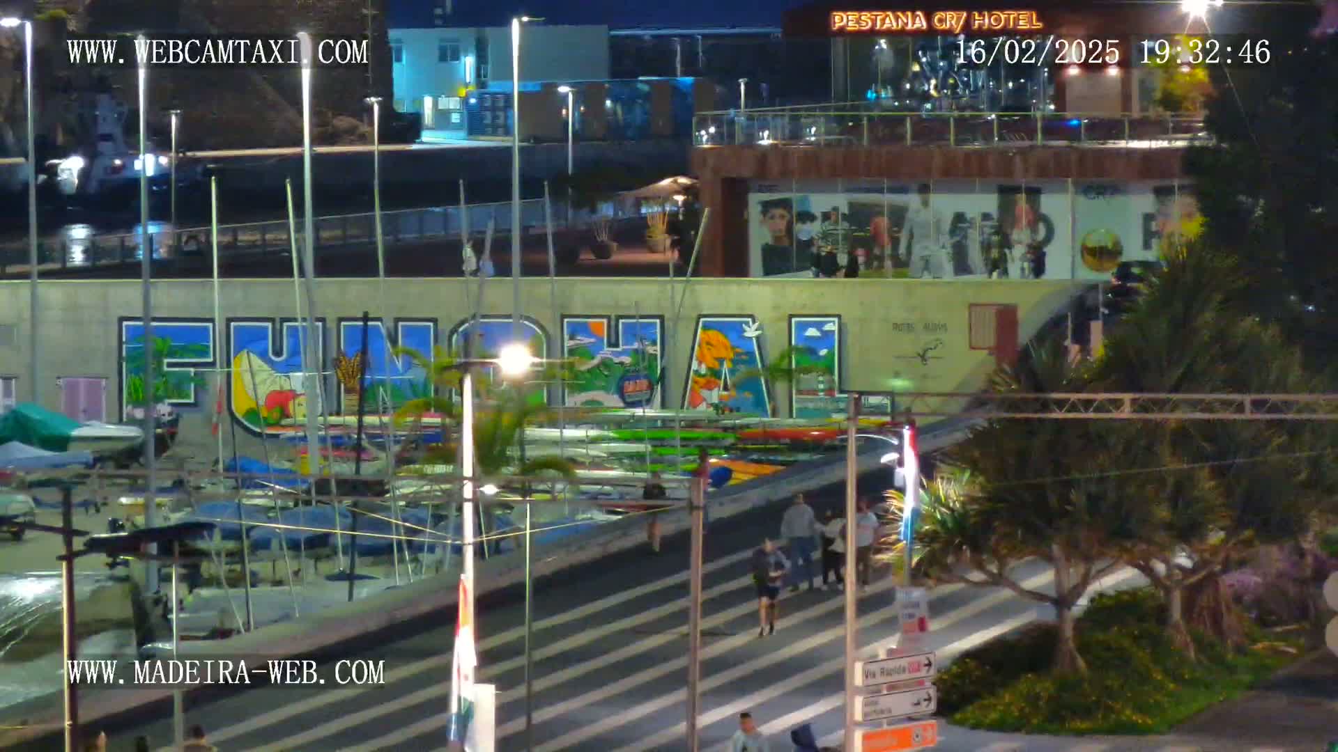 A nighttime view of a marina with numerous boats, a large mural reading "Funchal," and pedestrians walking along a walkway near palm trees.