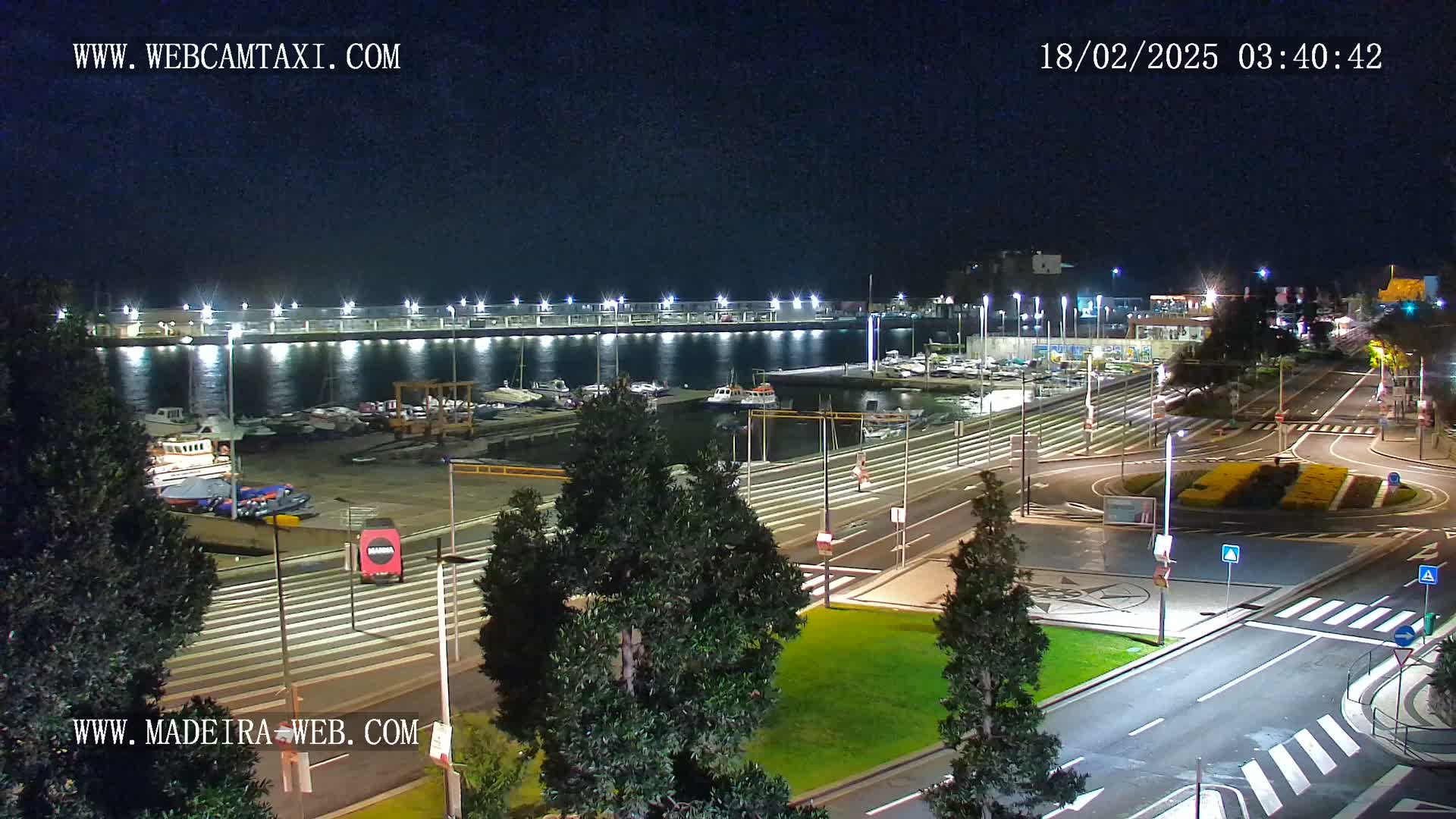 A nighttime, illuminated view of a harbor filled with boats, adjacent to a roadway with traffic circles and green spaces.