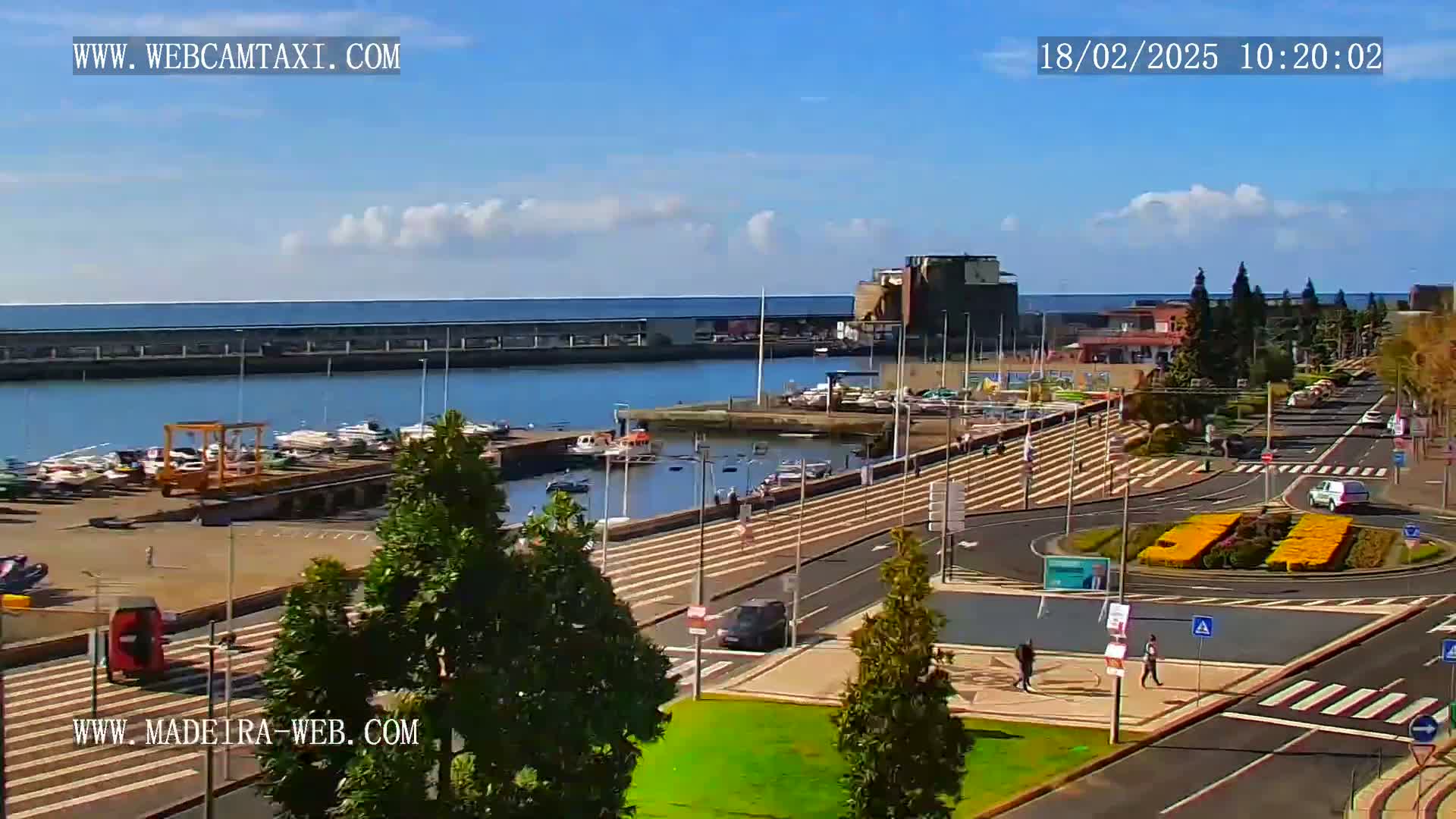 A marina with numerous boats is visible from a high vantage point on a partly sunny day, overlooking a paved road with a roundabout and sparse pedestrian traffic.