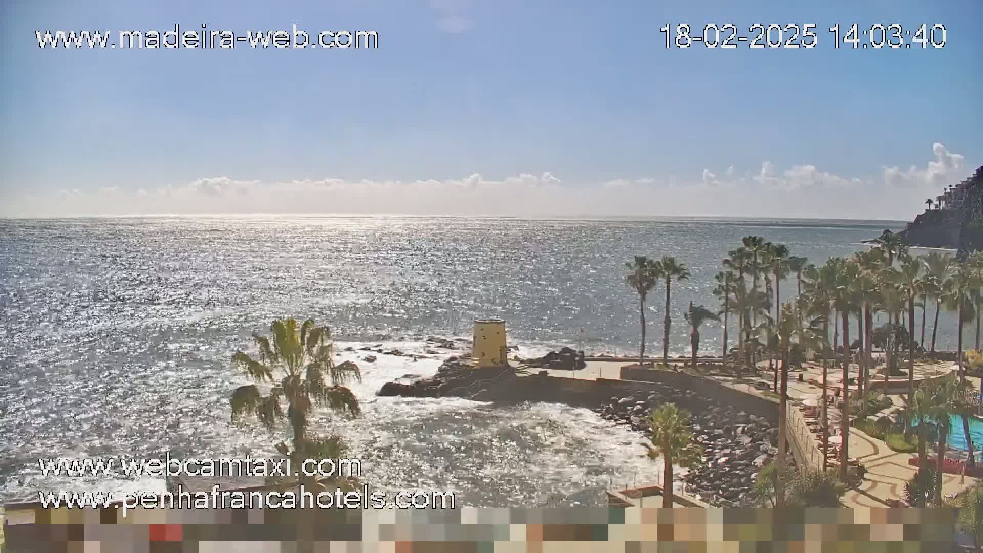 A partly sunny day shows a rocky coastline with palm trees and a small tower, overlooking a wavy ocean.