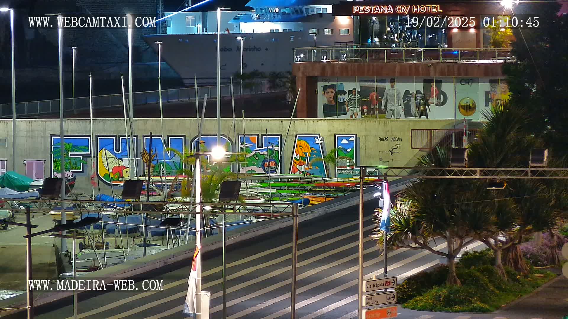 A nighttime harbor scene shows a large mural of "Funchal" on a wall behind a marina filled with boats, illuminated by streetlights under a clear sky.