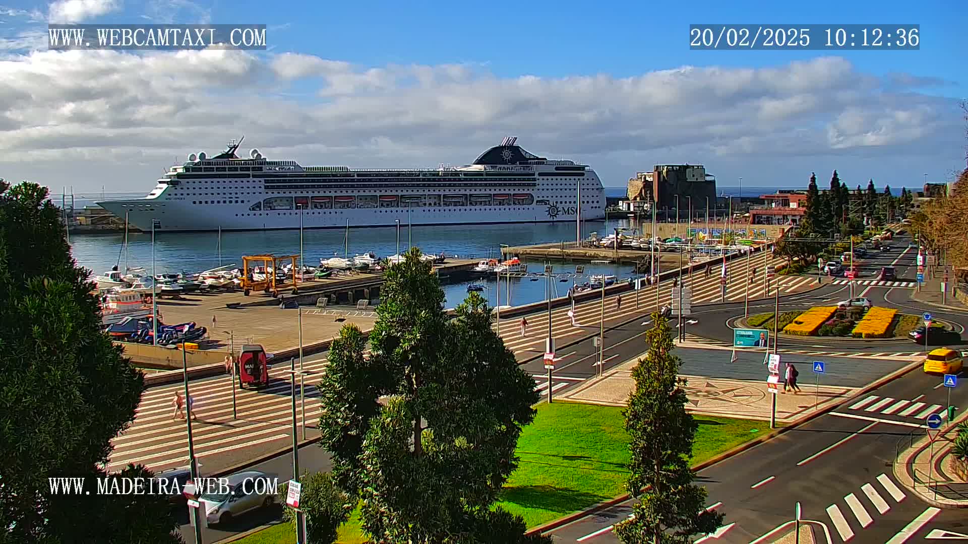 A large cruise ship is docked in a harbor, overlooking a town square with pedestrian walkways and several cars under a mostly sunny sky with some clouds.