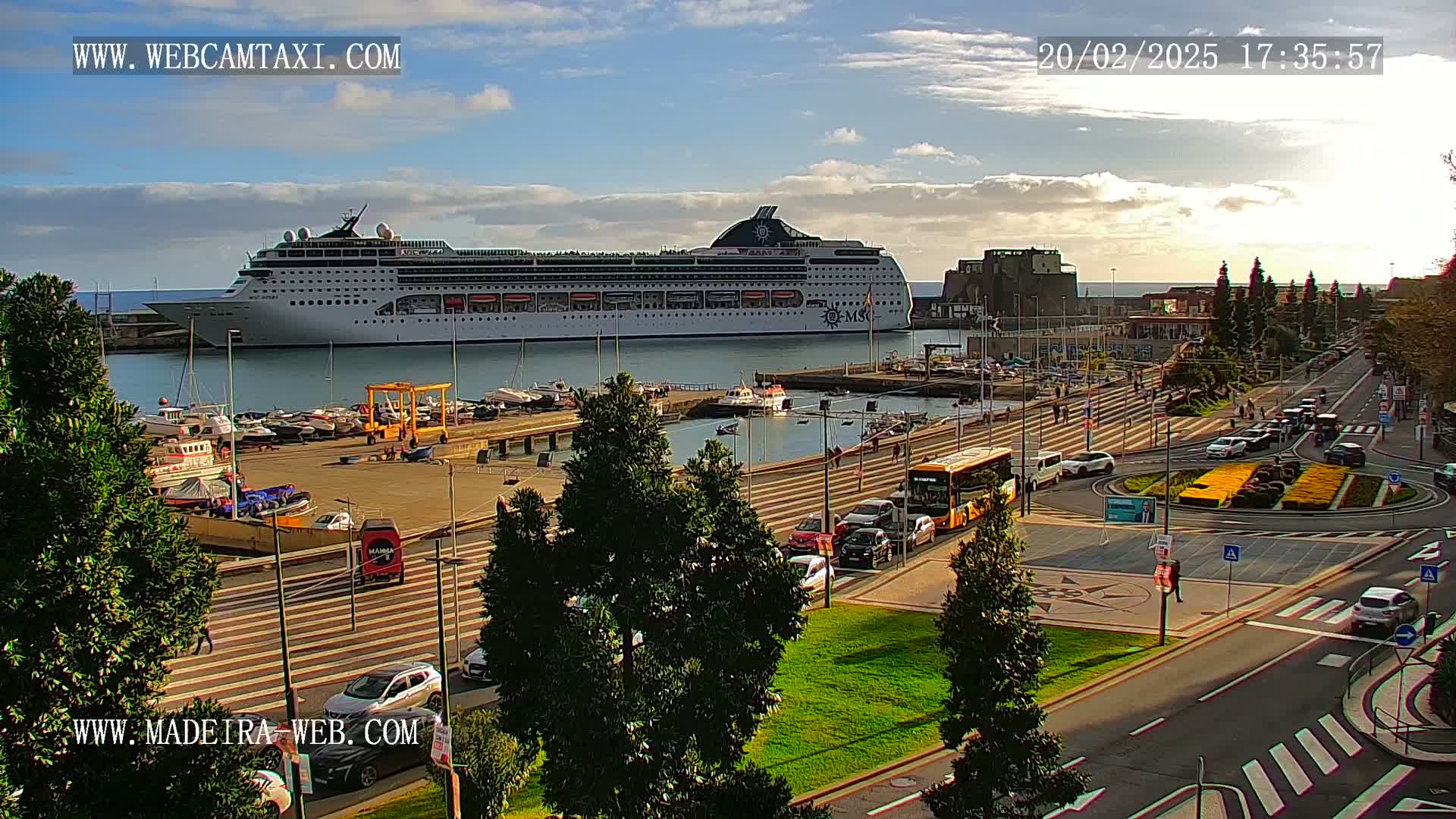 A large cruise ship is docked in a harbor under a partly cloudy sky, with cars and a bus traveling on a nearby road.