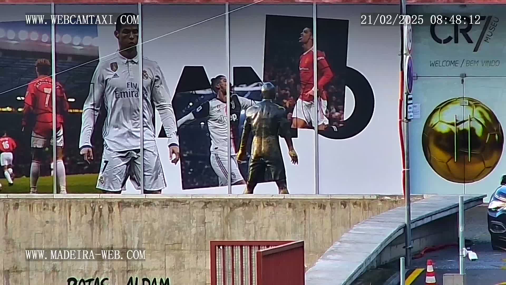 A building exterior displays large images of Cristiano Ronaldo in different soccer jerseys, a golden soccer ball, and a bronze-colored statue of a person, all under overcast skies.