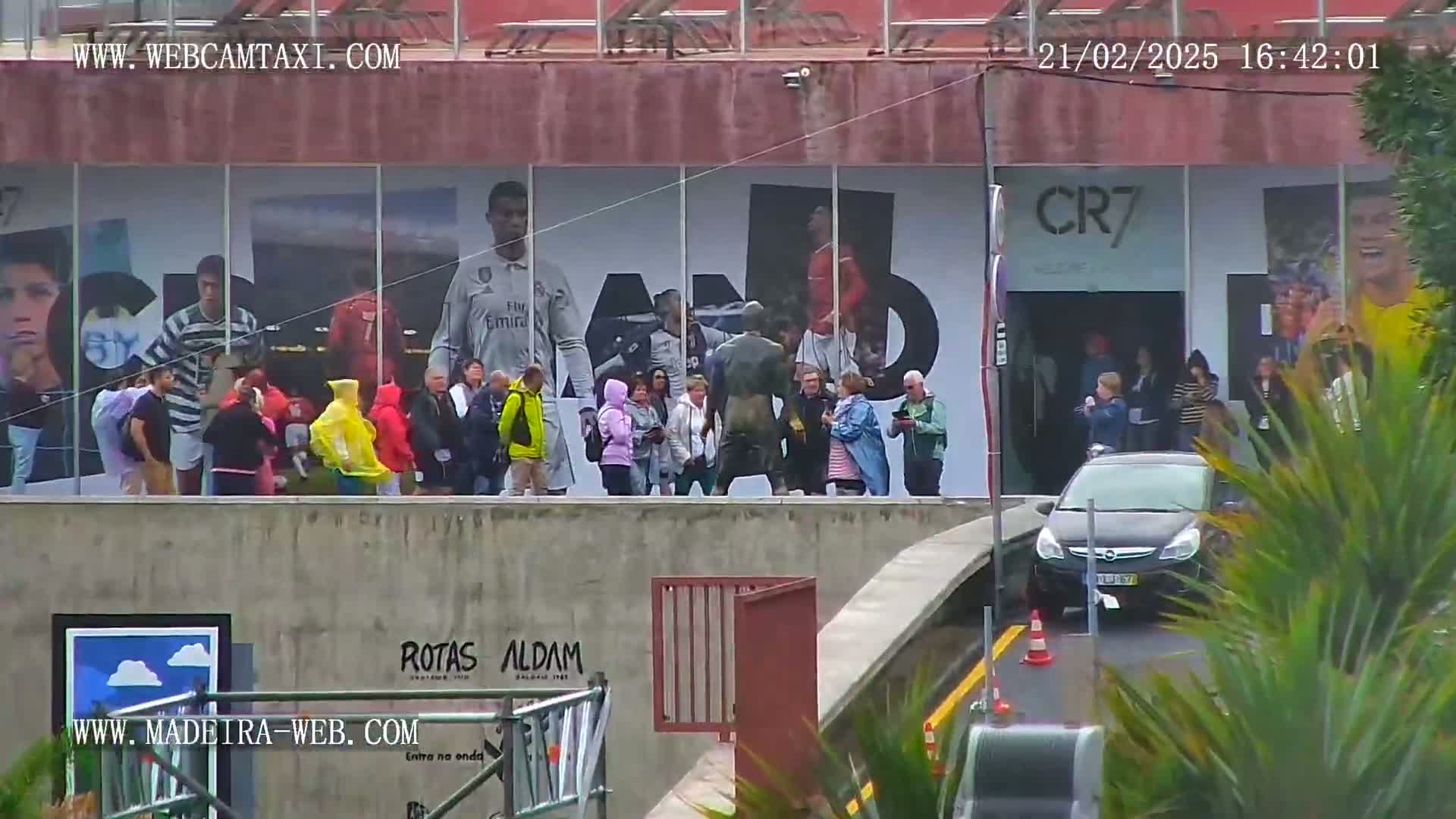 A group of people stand in front of a building with large images of soccer players on it, a car is parked nearby, and the weather appears to be overcast or rainy.