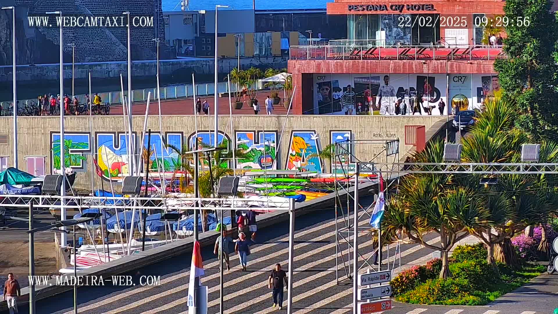 A sunny harbor scene features a large mural of the word "FUNCHAL," numerous boats docked, and pedestrians walking on a striped walkway.