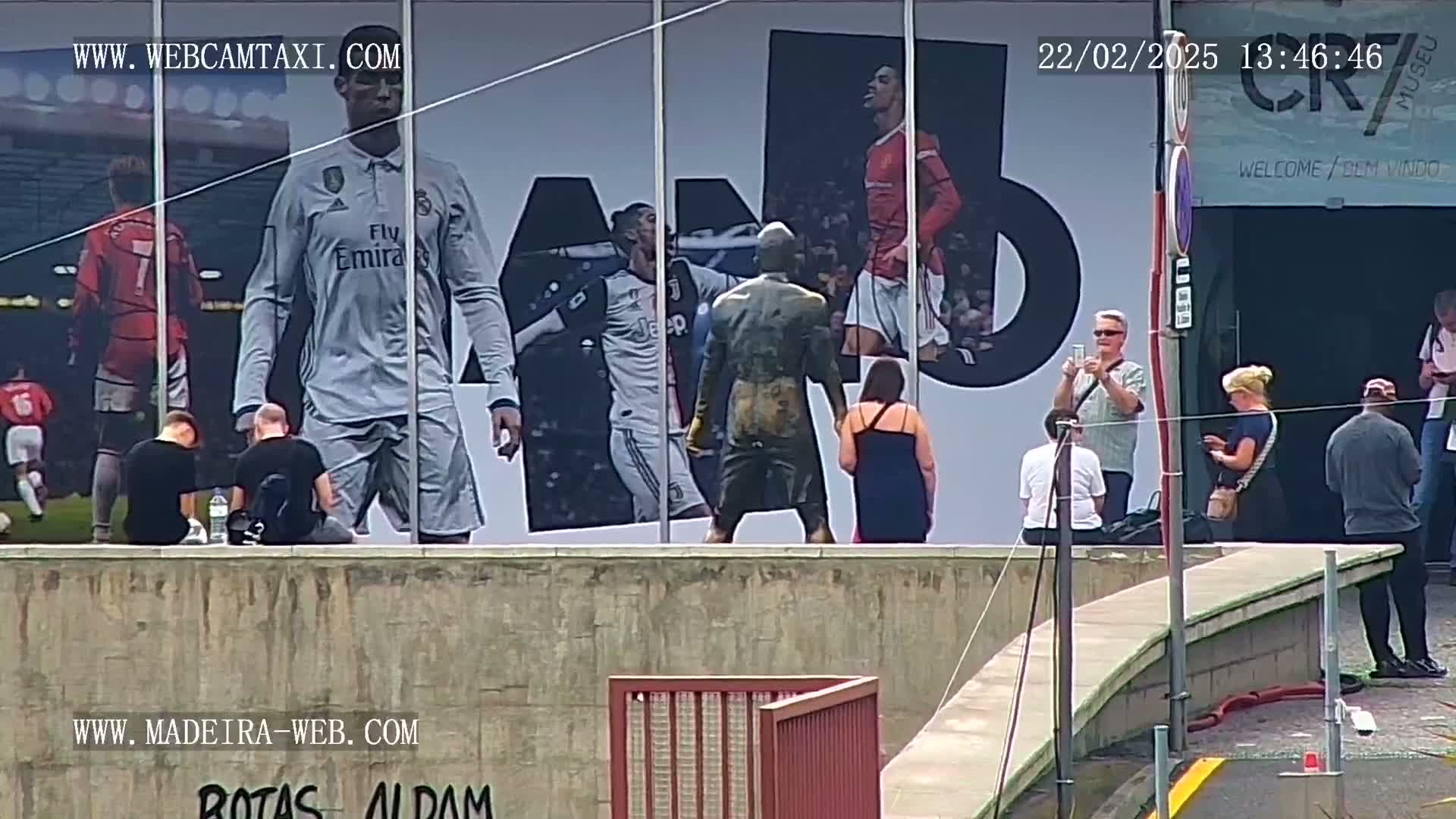 A large billboard depicting Cristiano Ronaldo in various soccer uniforms is viewed from across a low wall by several people, some of whom are taking pictures.