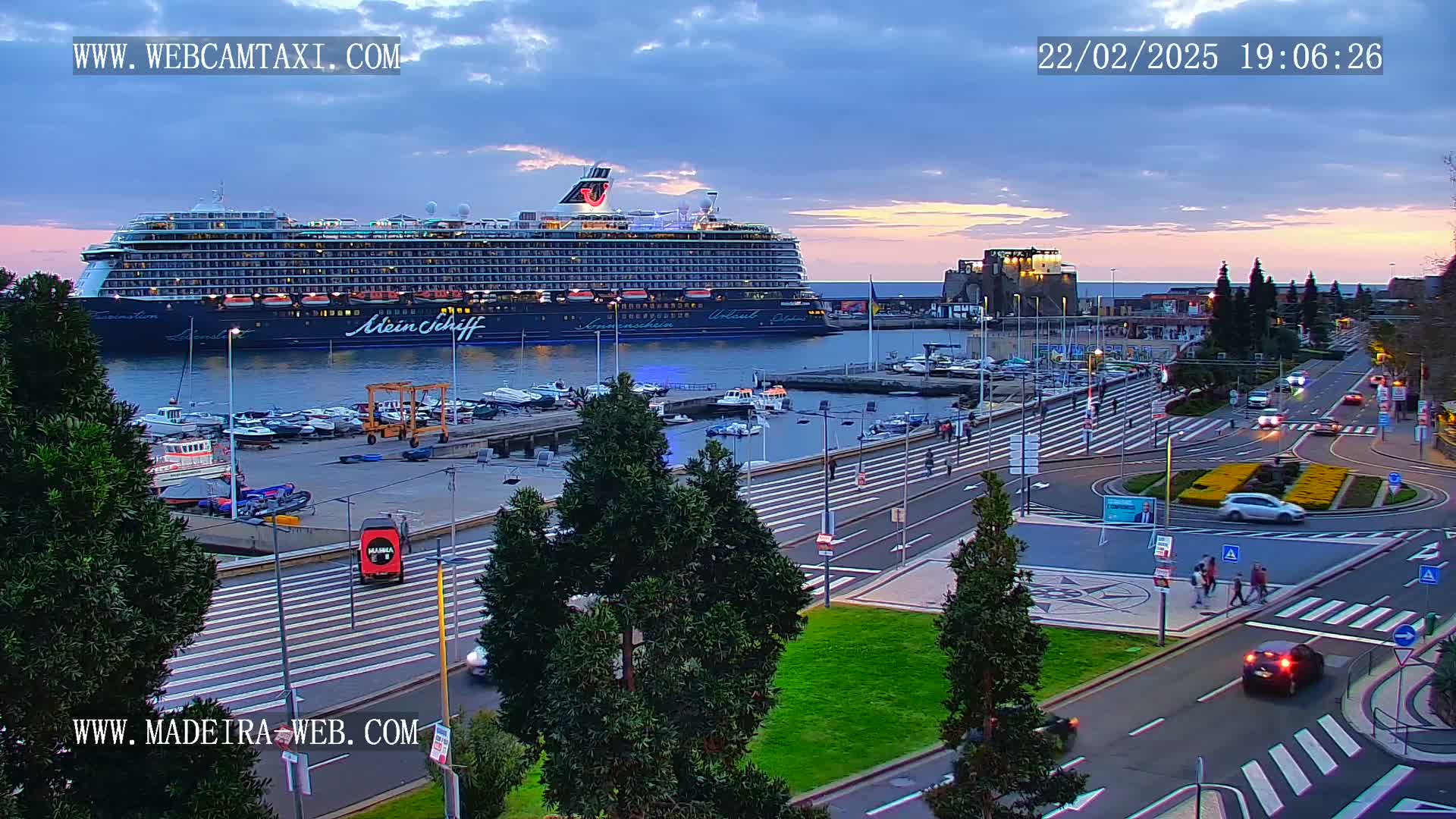 A large cruise ship is docked at a harbor during a twilight hour with a mostly clear sky, and the surrounding area shows city streets and pedestrian crossings.
