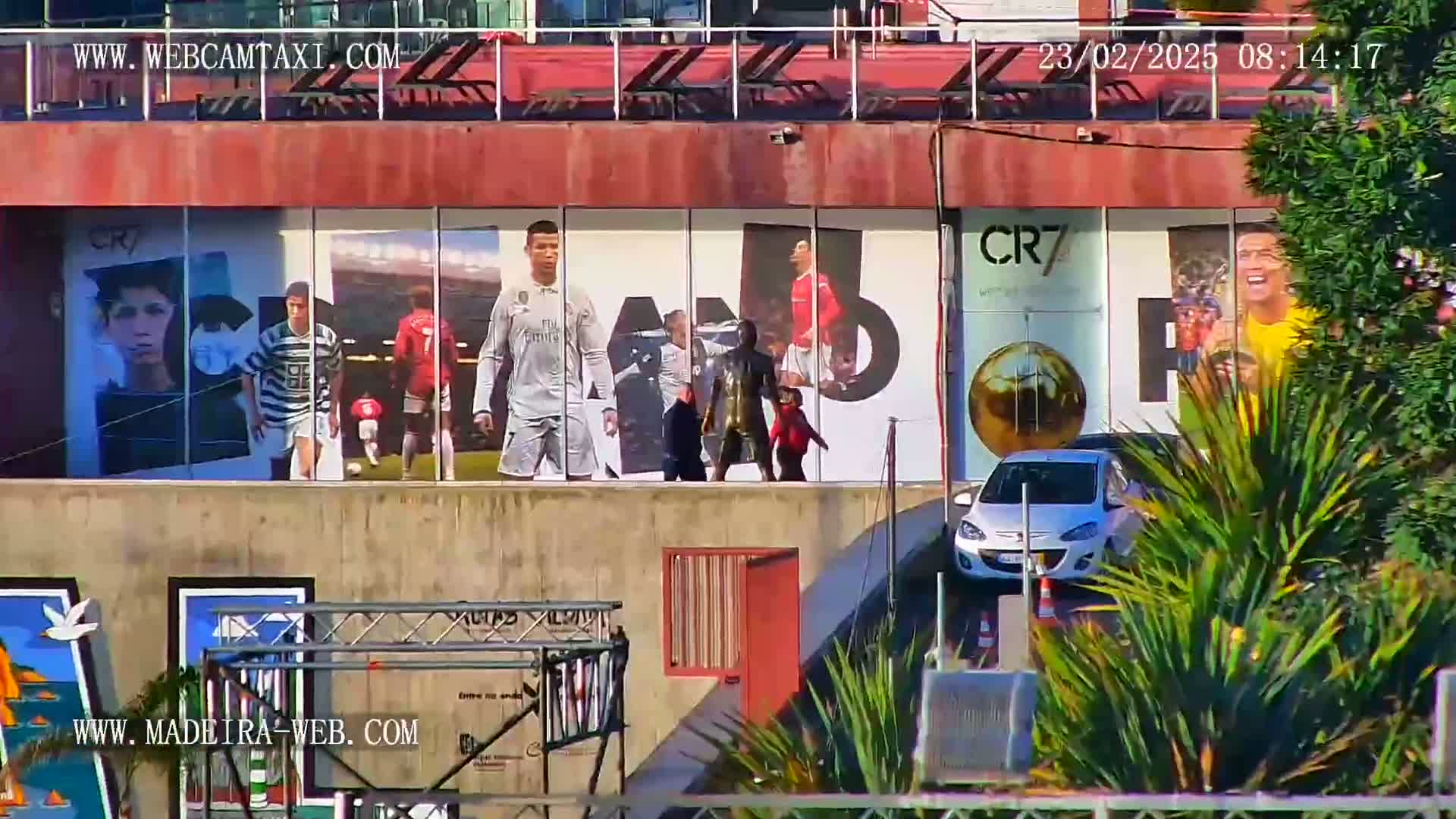 A building with large images of Cristiano Ronaldo and other soccer players is partially obscured by lush green plants, a silver car, and several people walking in front.