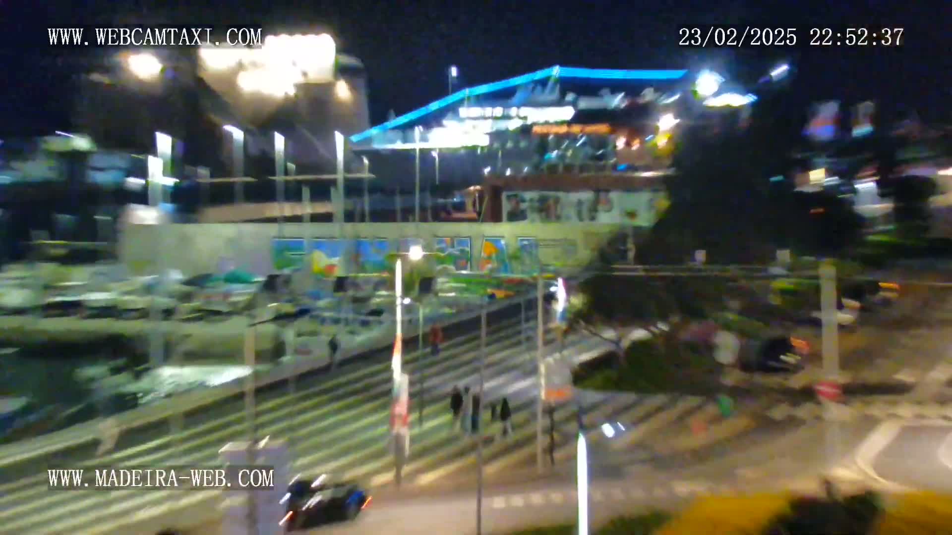 A nighttime, blurry view of a waterfront area with brightly lit buildings, parked vehicles, and pedestrians crossing a street.