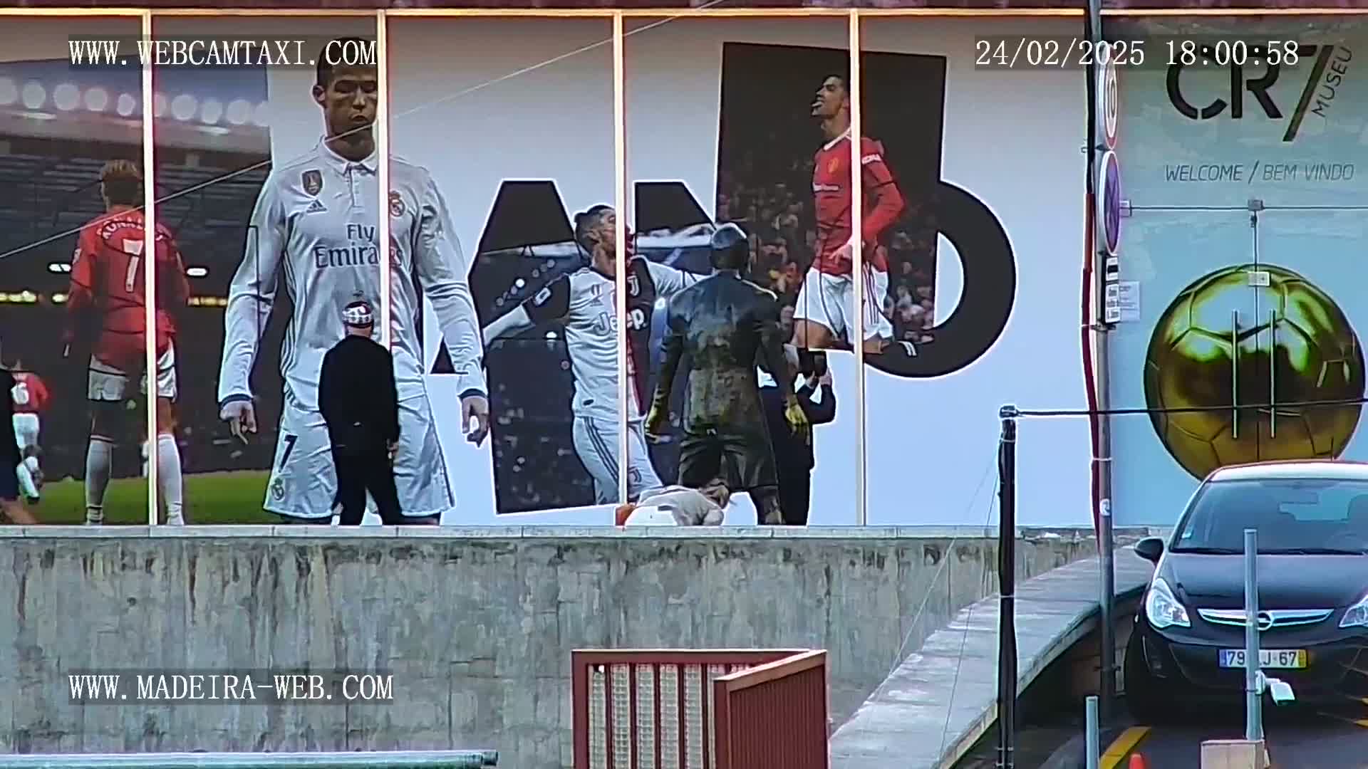 A large outdoor mural depicting Cristiano Ronaldo in various soccer jerseys is visible, with a black car parked in front of it and overcast skies.
