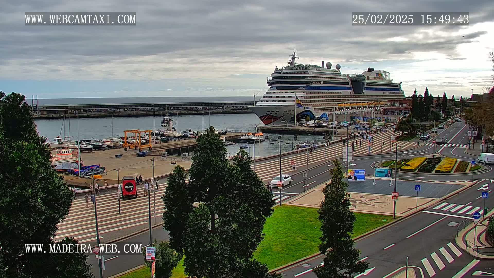 A large cruise ship is docked at a harbor on an overcast day, with a paved area and green spaces visible in the foreground.