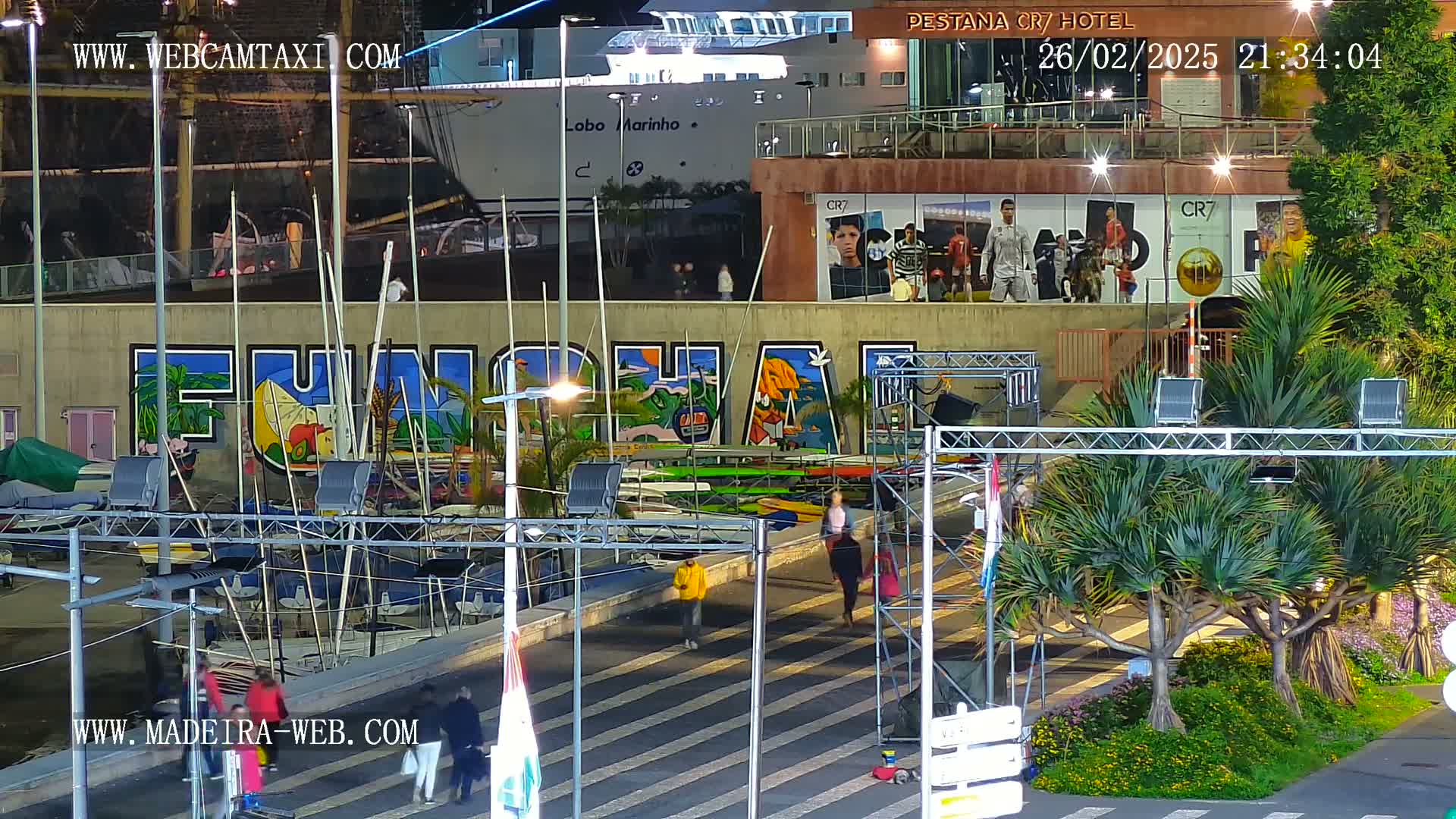 A nighttime harbor scene shows a large mural of the word "FUNCHAL" painted on a wall, with boats moored alongside and people walking on a nearby roadway under bright lights.