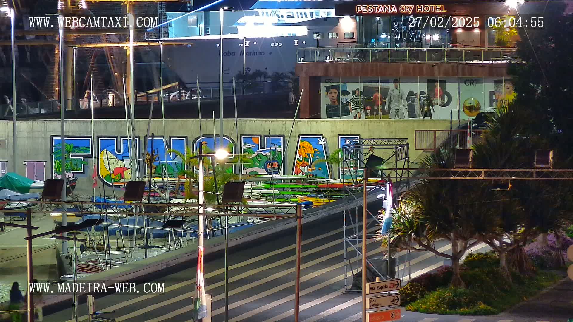 A nighttime view of a harbor with numerous sailboats docked, a large mural depicting the word "FUNCHAL," and a building displaying illuminated advertisements.