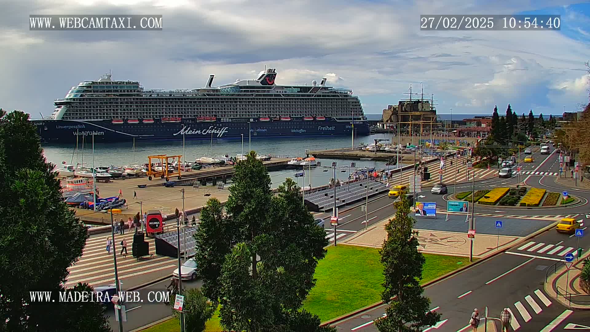 A large cruise ship is docked at a harbor on a partly cloudy day, with cars and people visible on the streets and sidewalks nearby.