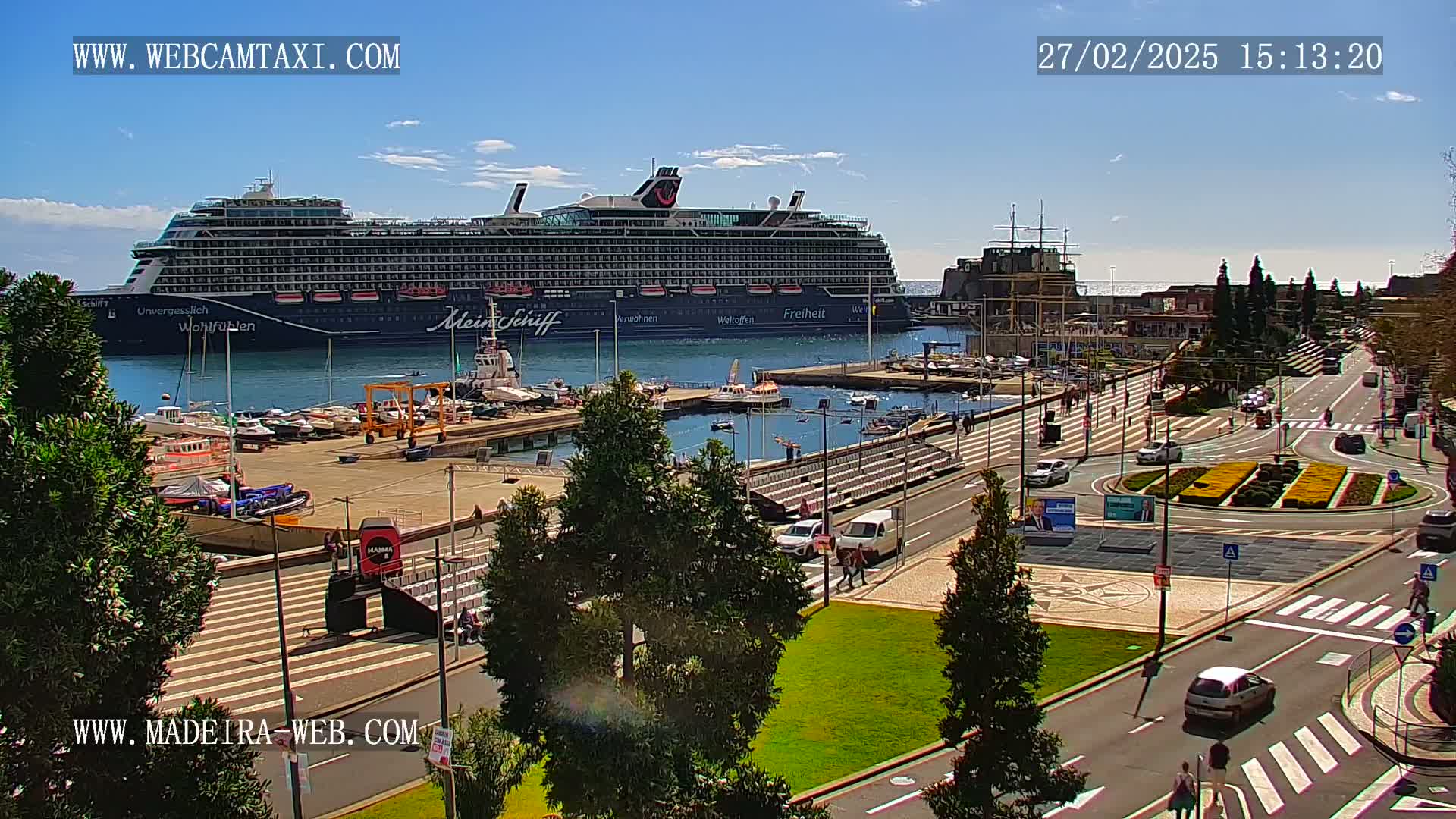 A large cruise ship is docked at a harbor on a sunny day, with cars driving along a waterfront road and pedestrians walking on sidewalks.