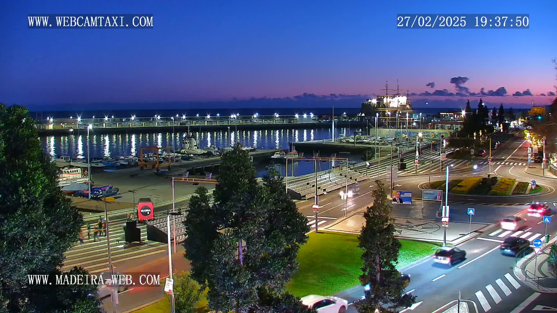 A marina at twilight, with cars driving on a road next to a grassy area, under a mostly clear sky.