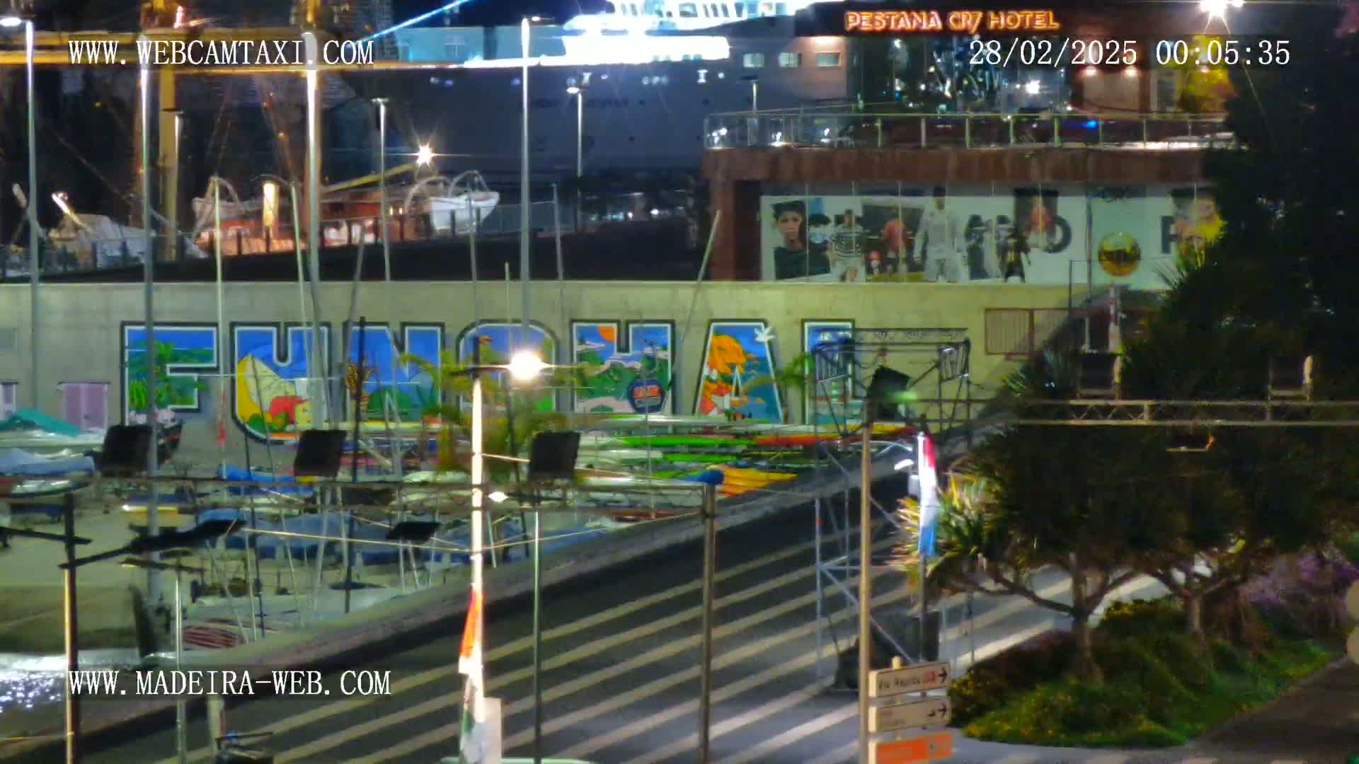 A nighttime view of a marina with numerous boats and kayaks, a large mural depicting the word "Funchal," and a building with advertisements.