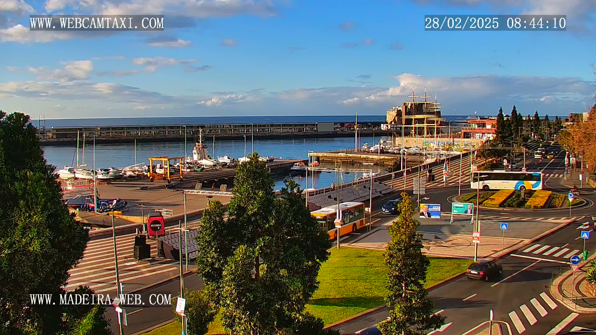 A harbor filled with boats is visible under a mostly sunny sky, with a city and roundabout in the foreground.