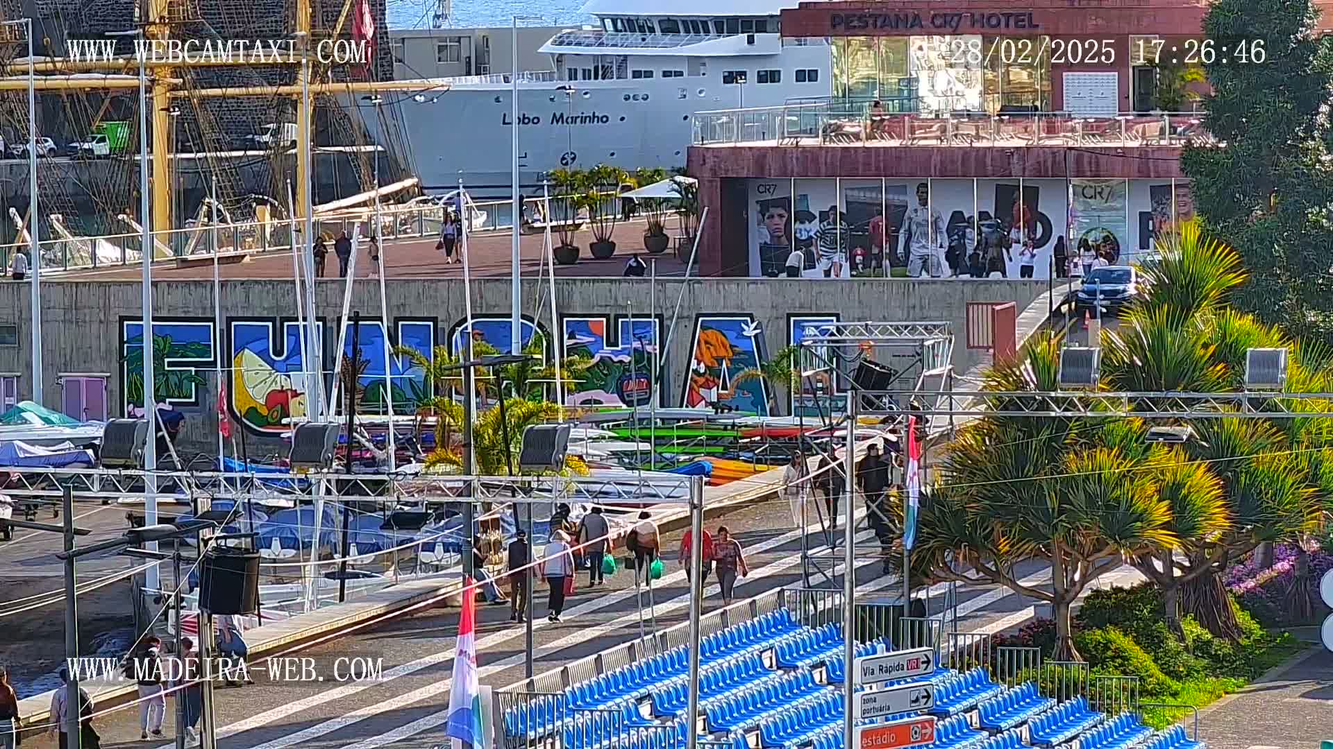 A sunny harbor scene shows a large mural, numerous boats, a cruise ship, and people walking along a paved area near rows of empty blue stadium seats.