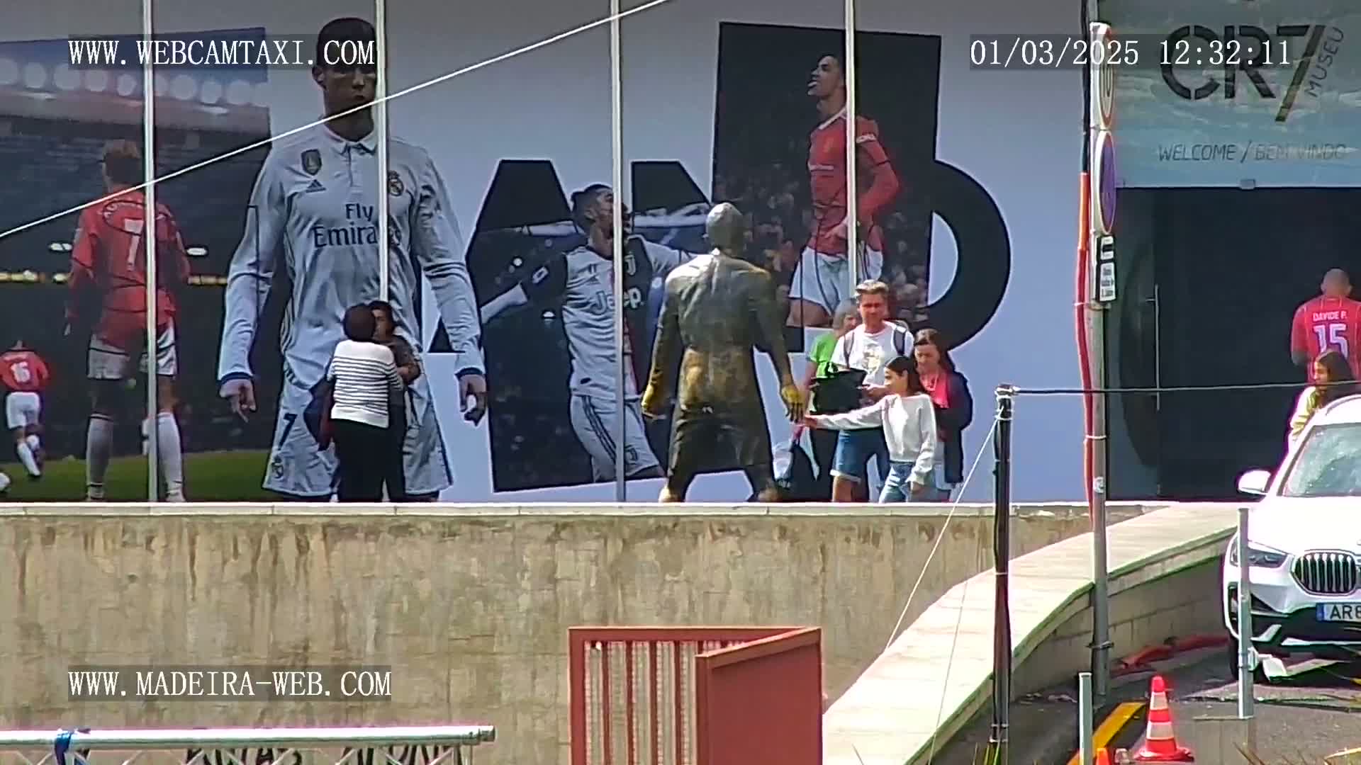 A large building features a mural of soccer players, a statue, and several people standing in front of it on a sunny day.