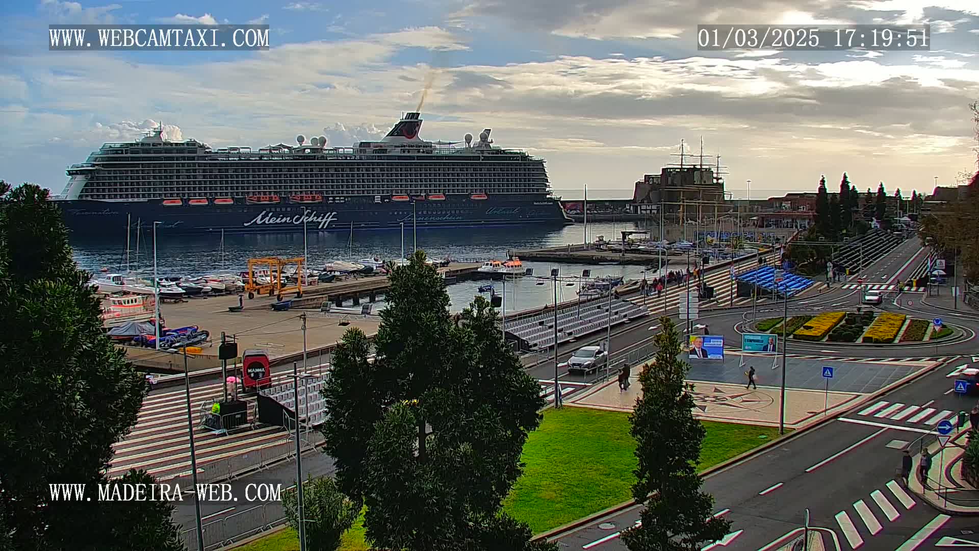 A large cruise ship is docked at a harbor, under a partly cloudy sky, near a city with a roundabout and several cars.