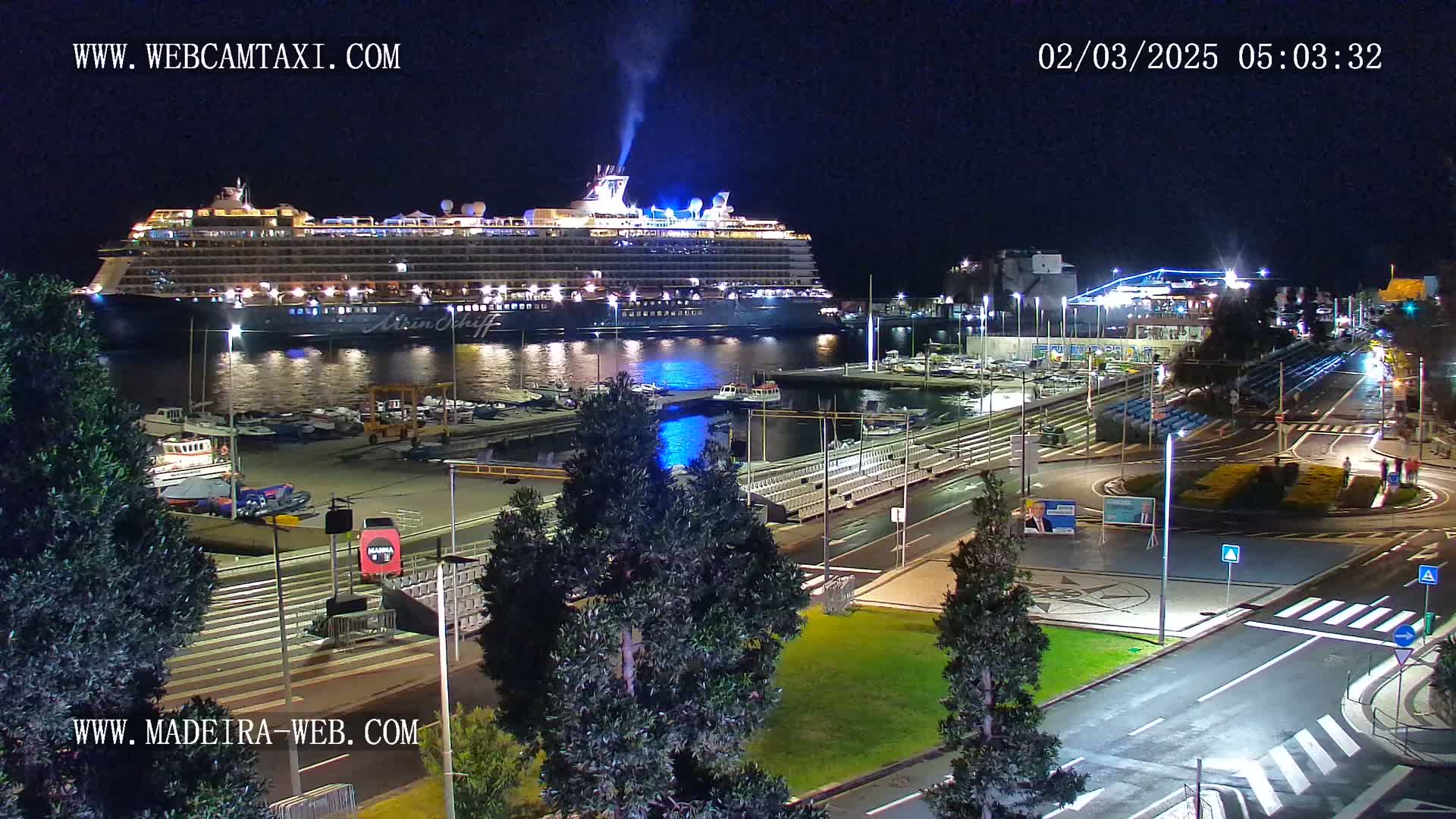 A large cruise ship is docked at a harbor at night, with surrounding streets and small boats visible.