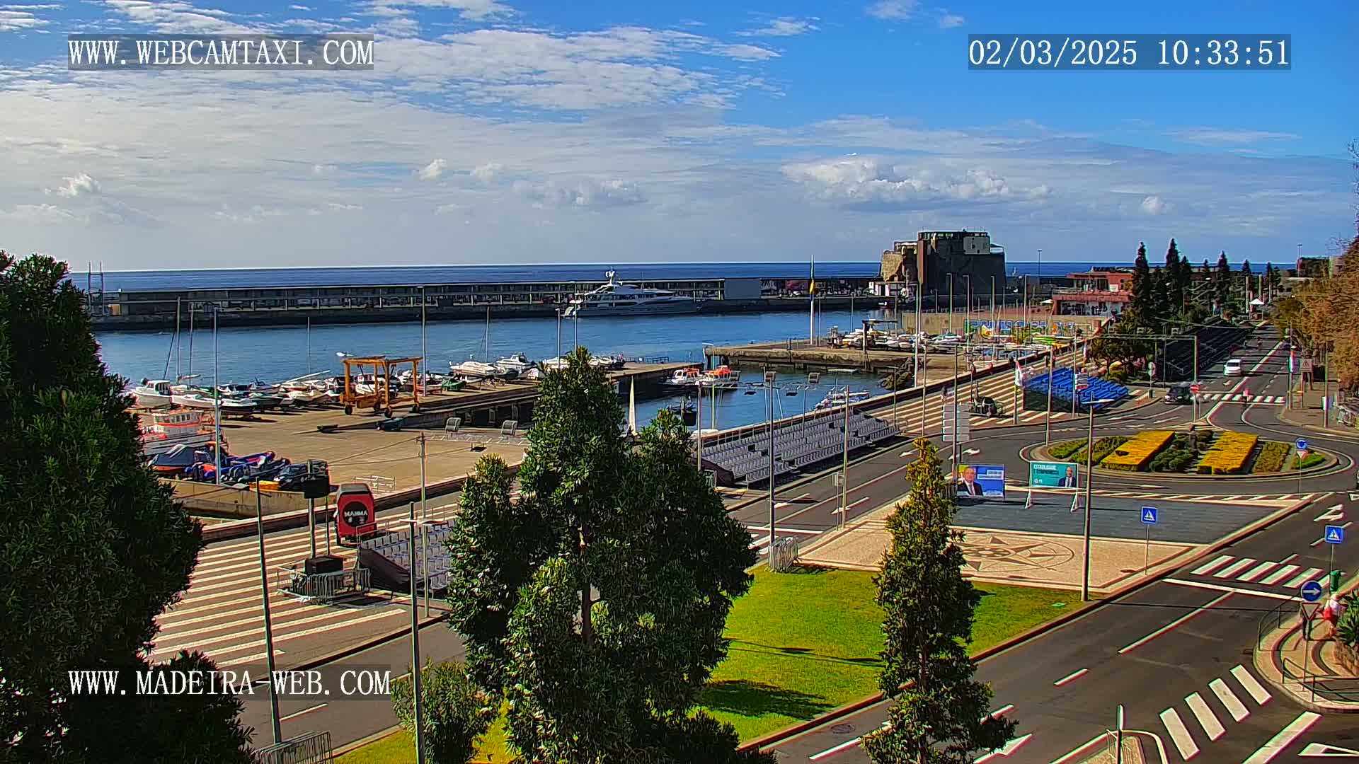A harbor filled with boats sits under a partly cloudy sky, next to a paved area with landscaping and empty seating, all overlooking the ocean.