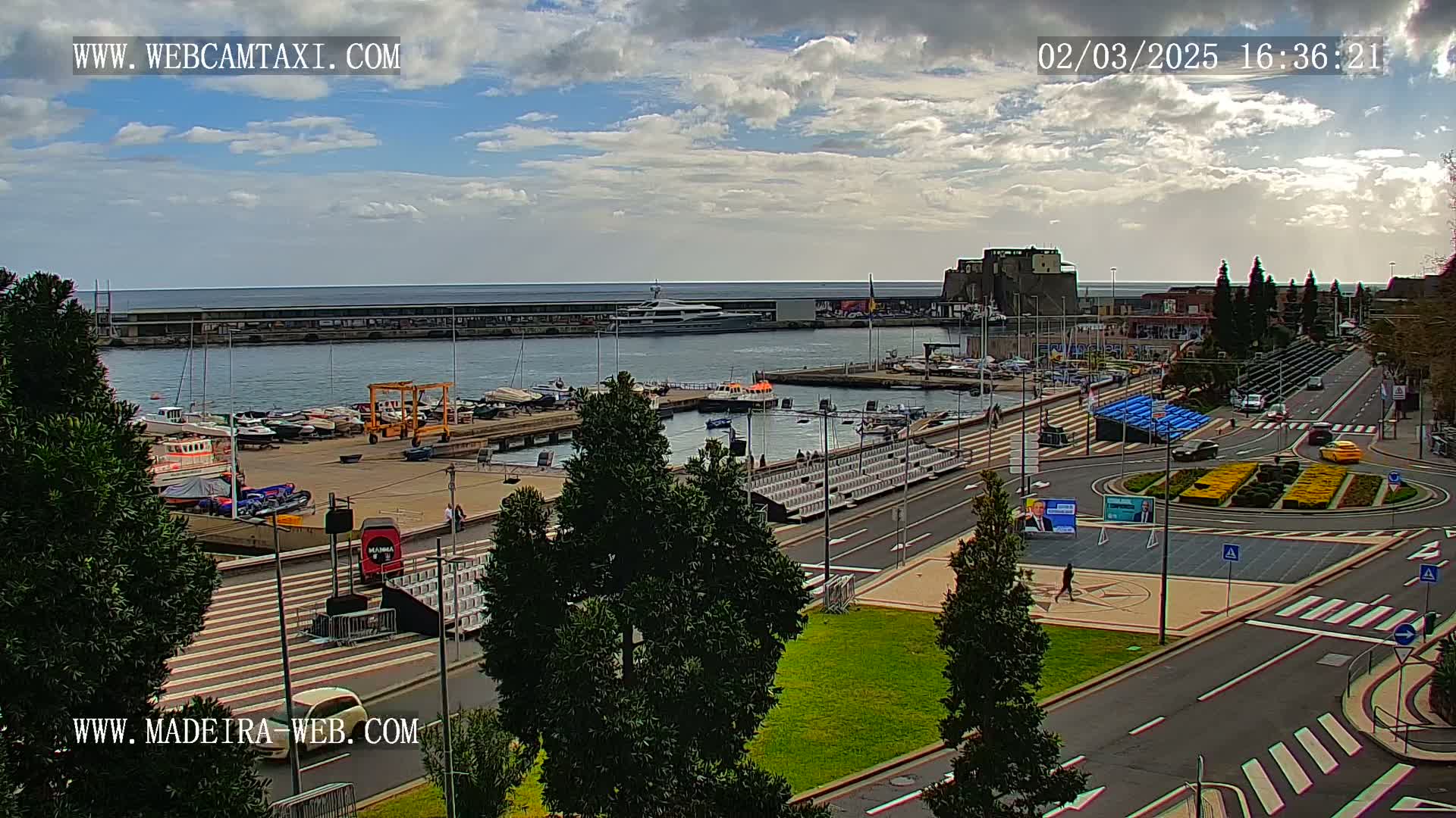 A partly cloudy day overlooking a harbor filled with boats and yachts, a city street with roundabouts, and green spaces.