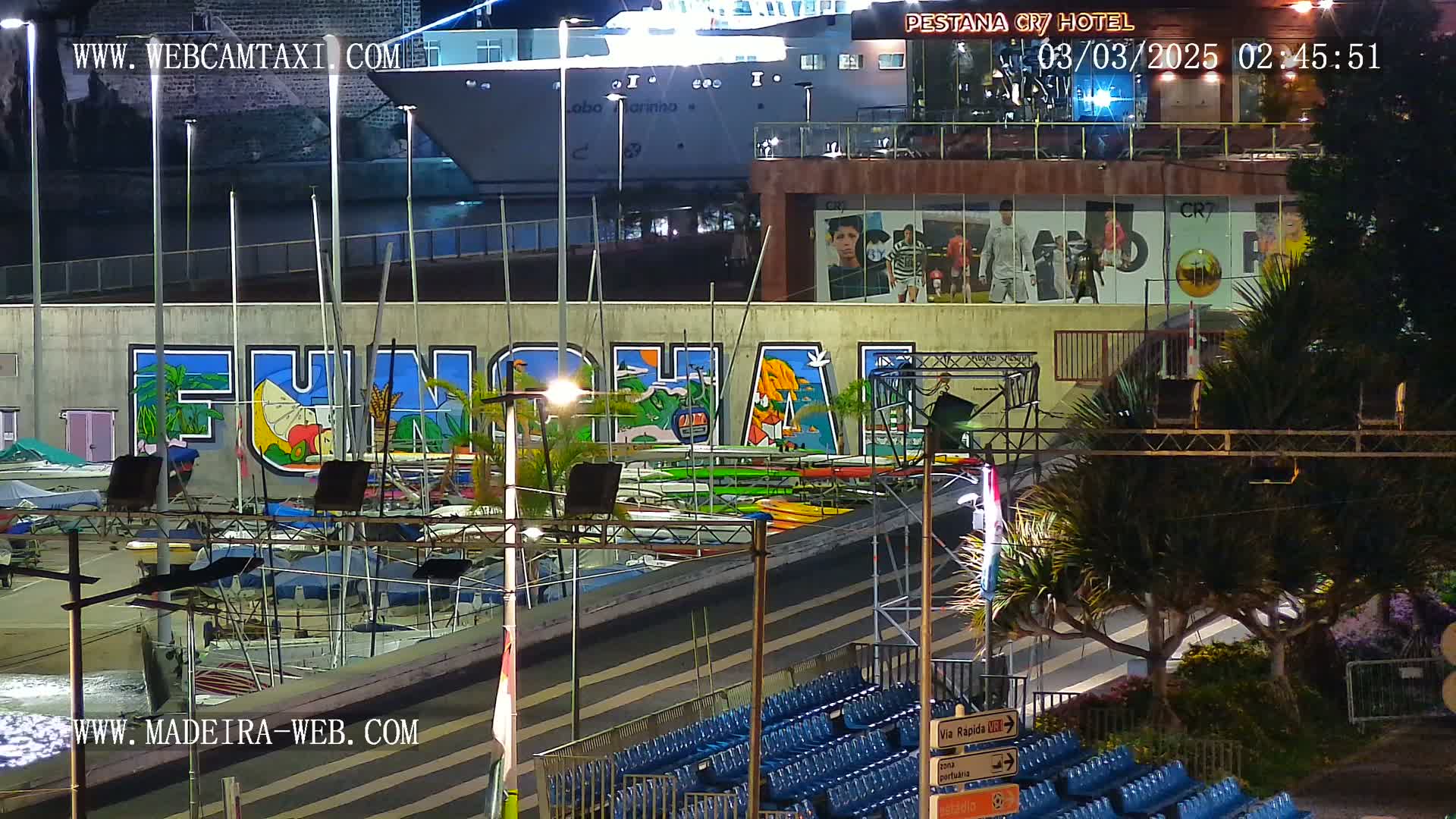 A nighttime view of a harbor with numerous boats docked, a large mural depicting the word "FUNCHAL," and a building with advertisements.