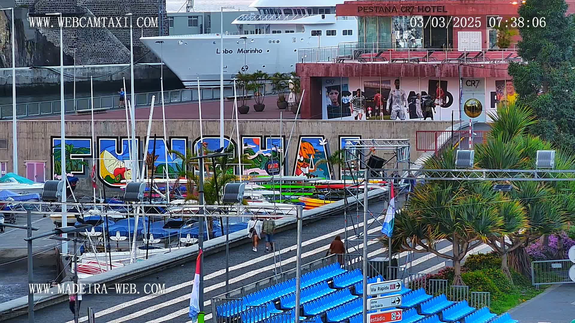 A harbor scene with a large mural, numerous boats, a cruise ship, and empty blue stadium seating, under partly cloudy skies.