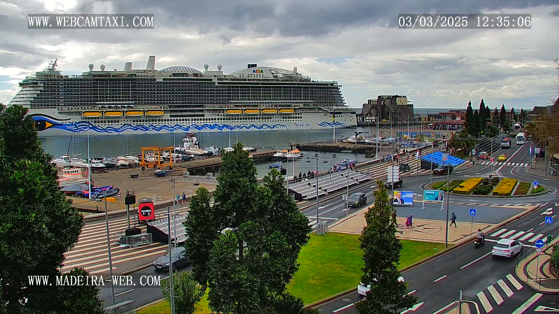 A large cruise ship is docked at a harbor under a cloudy sky, with cars and people visible on the surrounding streets and waterfront.