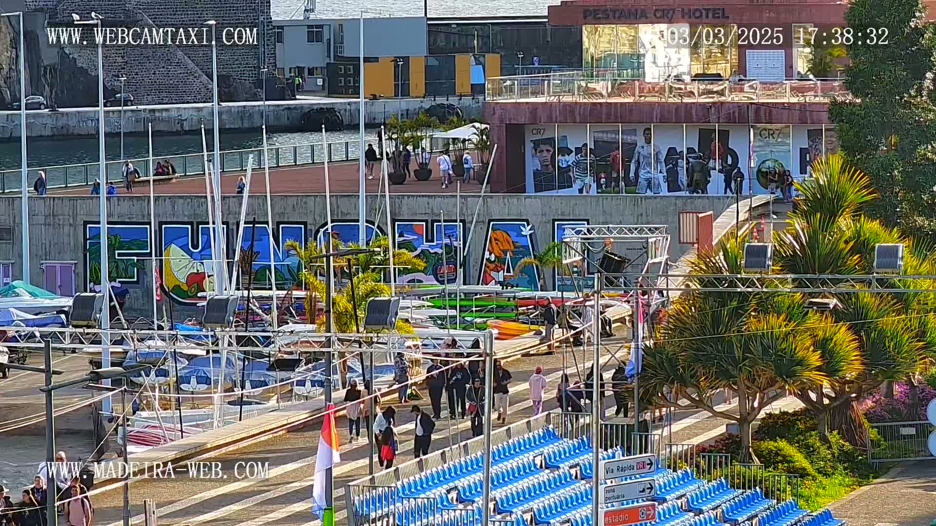 A sunny harbor scene shows numerous sailboats docked, a large mural, people walking, and empty blue stadium seating.