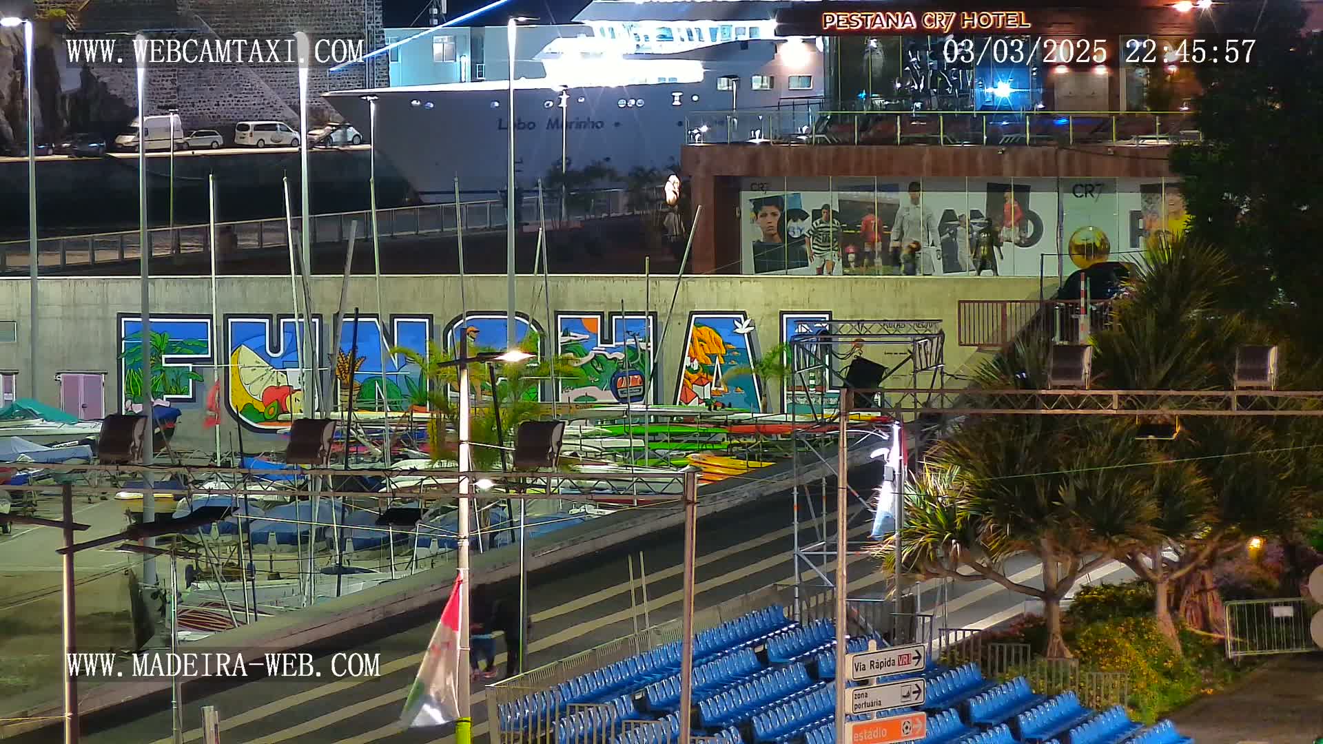 A nighttime harbor scene shows a large mural of the word "Funchal," boats docked below it, a cruise ship in the background, and empty blue stadium seating in the foreground under nighttime lighting.
