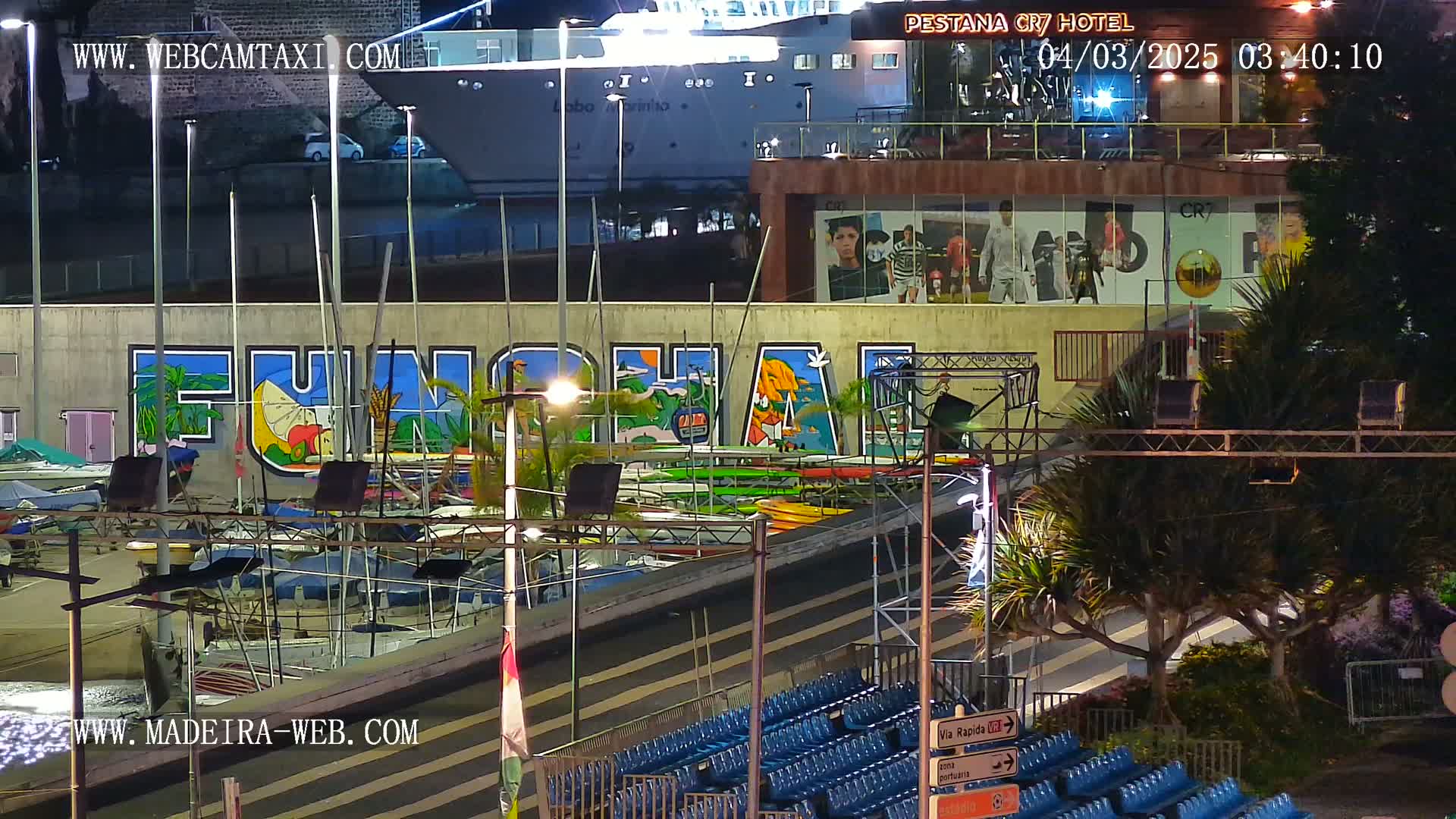 A nighttime harbor scene shows a large mural of the word "FUNCHAL" on a wall, behind a marina filled with boats, under artificial lights.