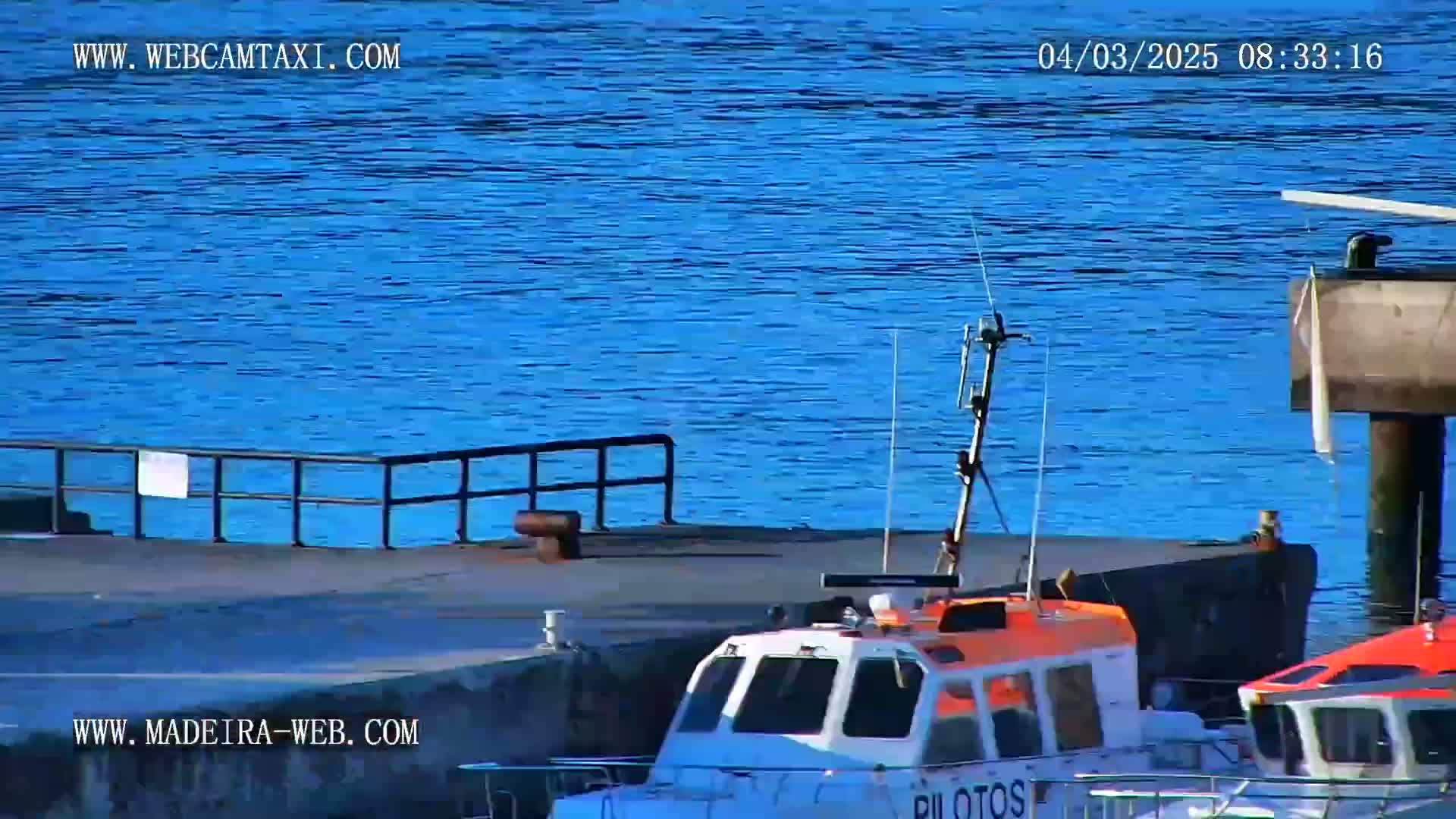 A small pilot boat is docked at a pier next to a larger, darker vessel, on a calm, blue body of water.