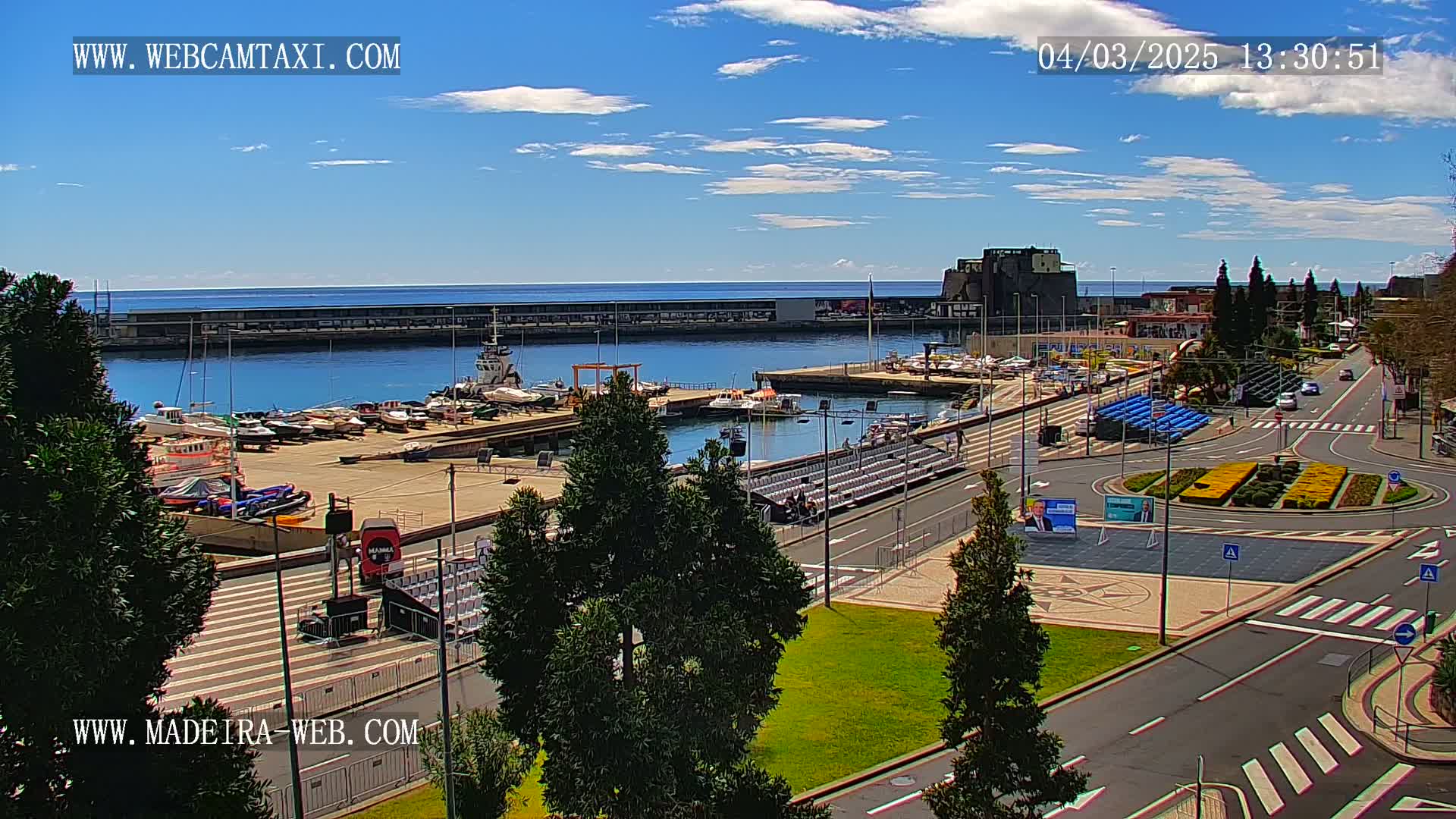 A marina filled with boats is seen under a mostly sunny sky, next to a paved area with temporary seating and a roundabout.