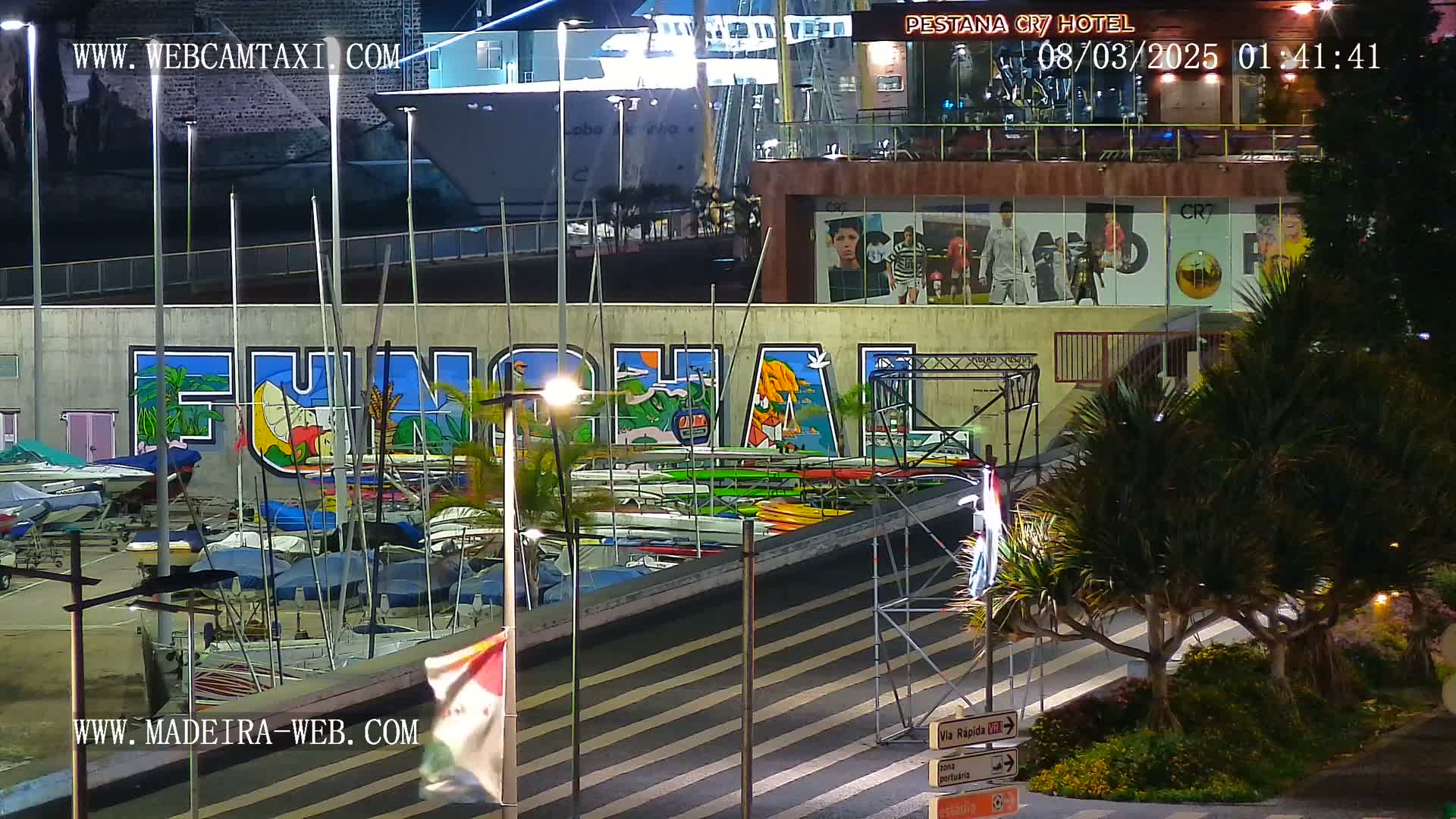 A nighttime harbor scene shows a large mural of "Funchal" on a wall behind numerous small boats and kayaks, with a large ship visible in the background under bright artificial lights.