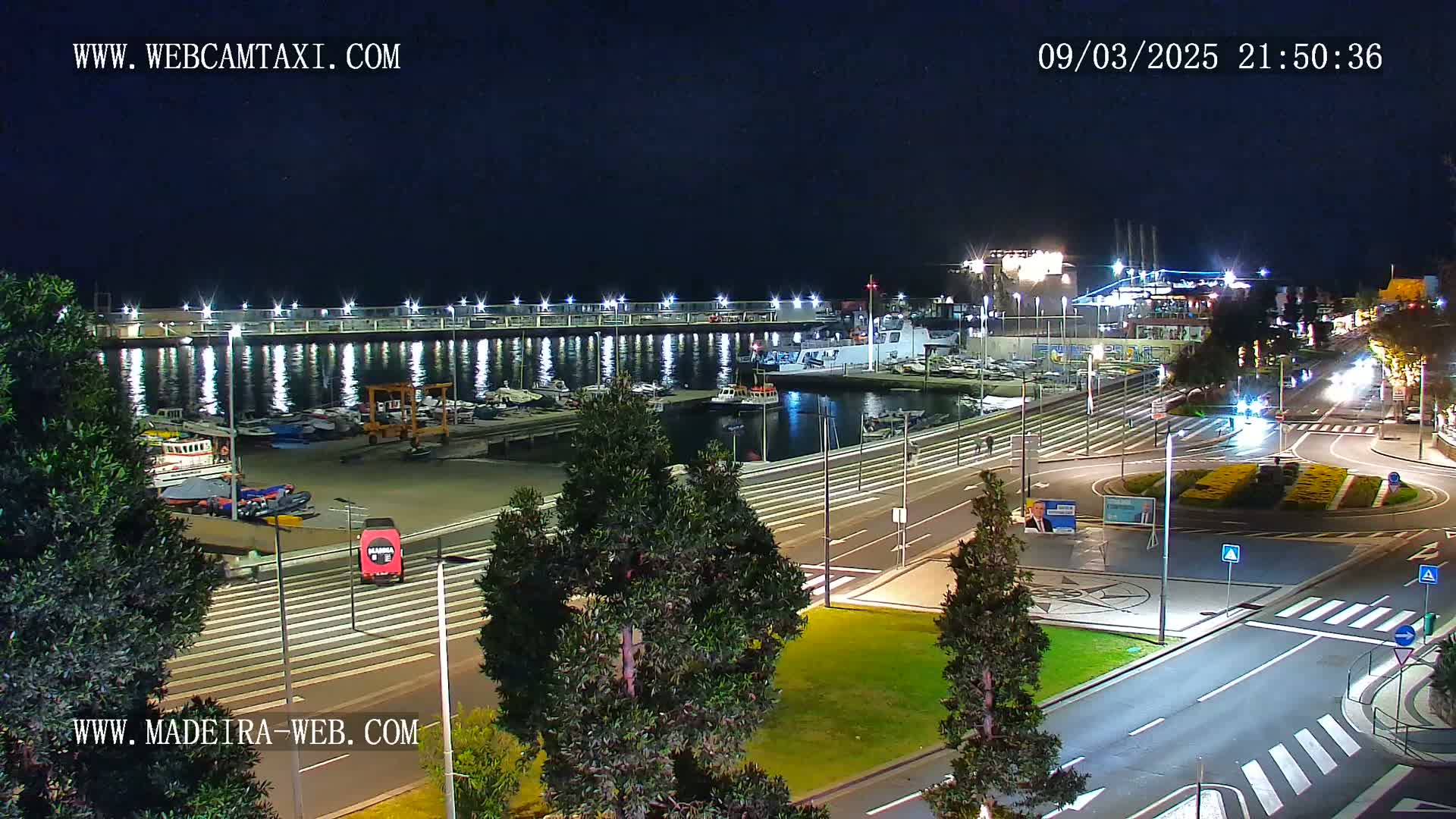 A nighttime view of a harbor filled with boats, adjacent to a roadway with minimal traffic under a clear, dark sky.