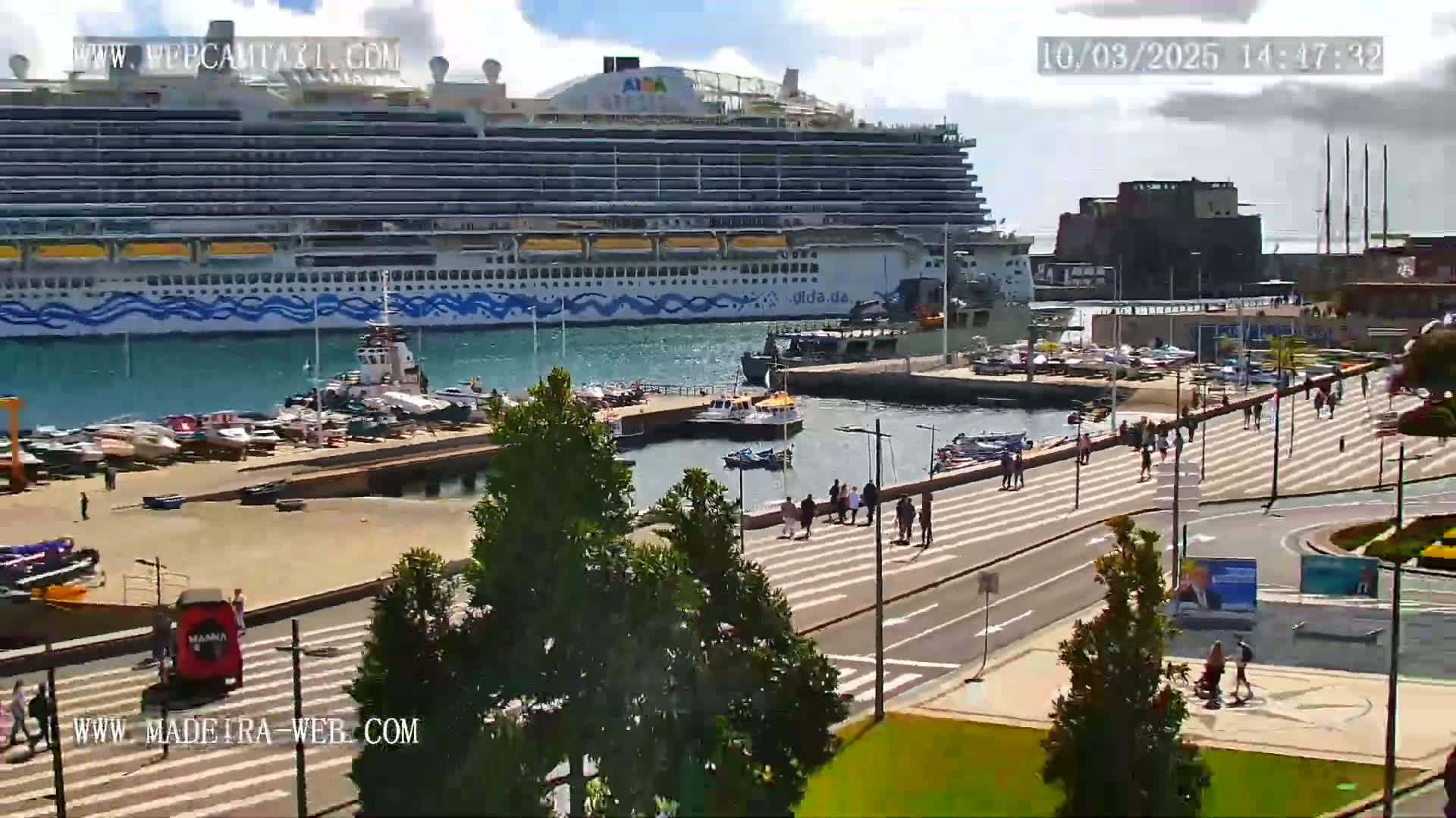A large cruise ship is docked at a harbor on a partly cloudy day, with several smaller boats and people walking along a nearby promenade.