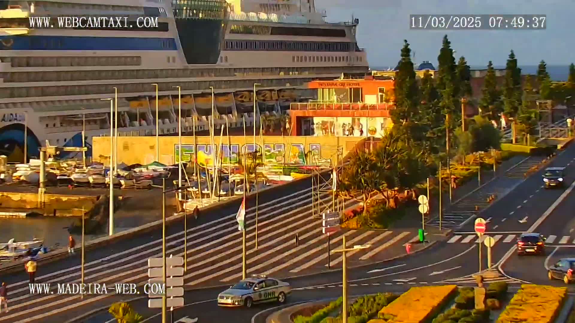 A large cruise ship is docked at a harbor next to a roadway with several cars, and pedestrians are walking along a waterfront promenade under sunny skies.